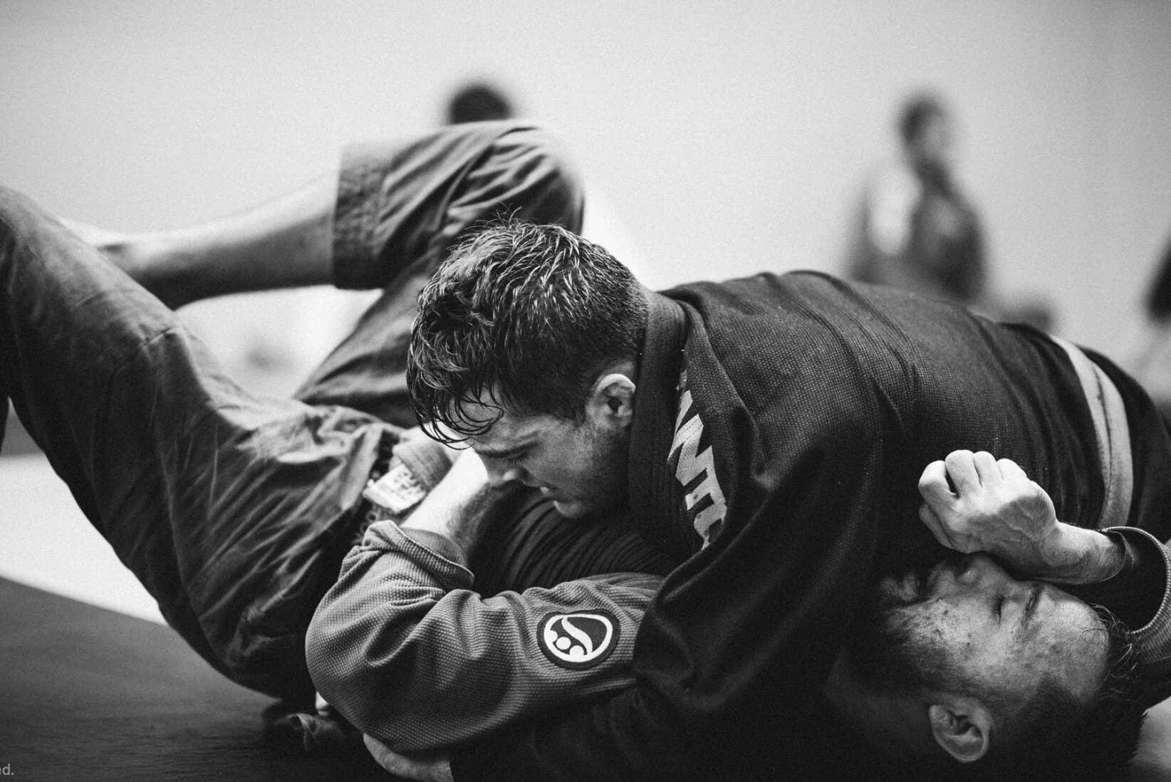 Black-and-white close-up of two athletes grappling on the mat, one in a dark gi working from top position, training partners blurred behind