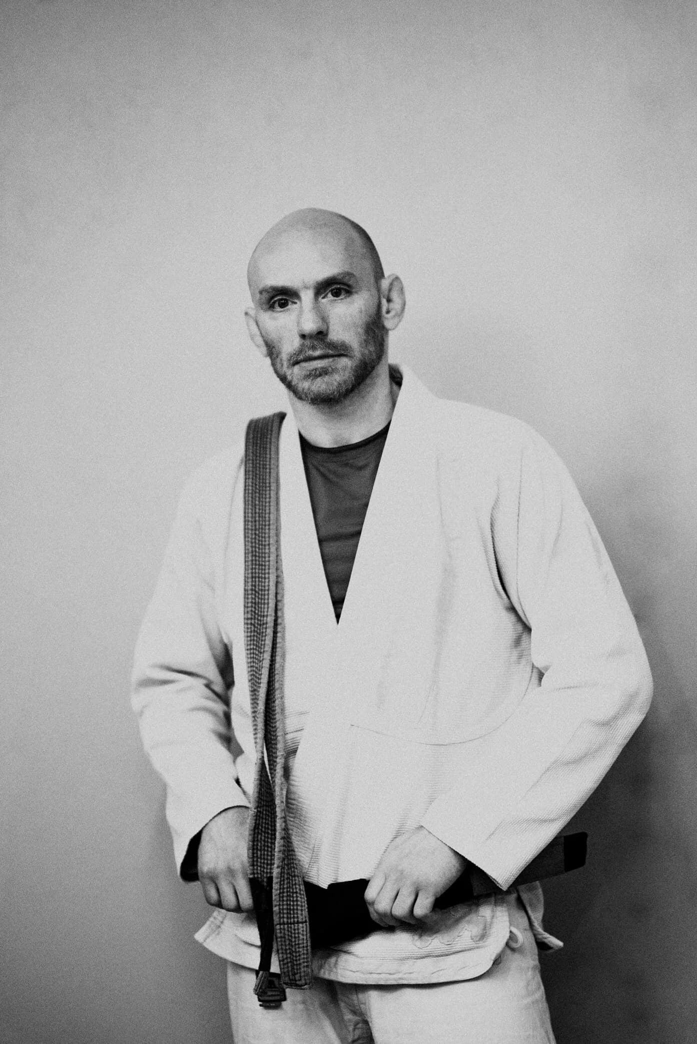 Black-and-white portrait of a bald, bearded athlete in a light gi fastening his black belt and looking into the camera, a second belt slung over his shoulder