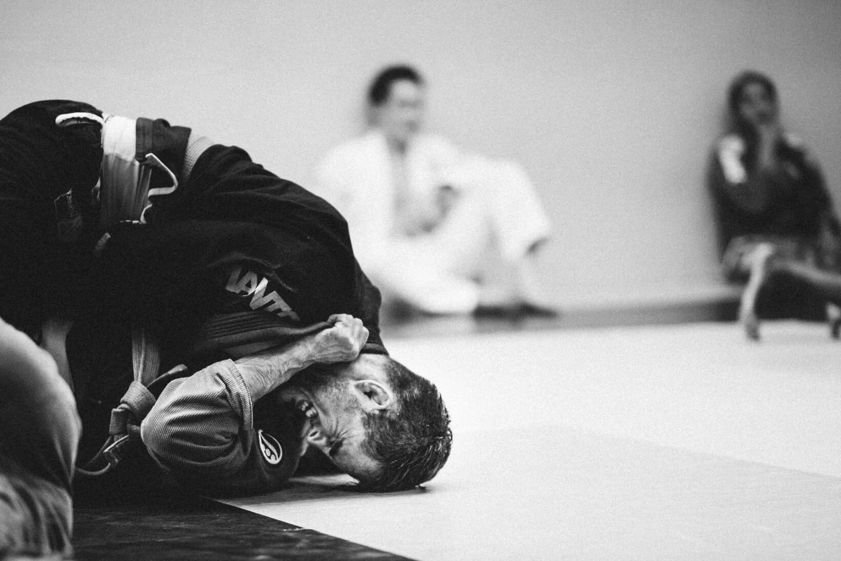 Black-and-white shot of an athlete face-down on the mat mid-roll, fist clenched in his own dark gi and grimacing, others watching from the wall
