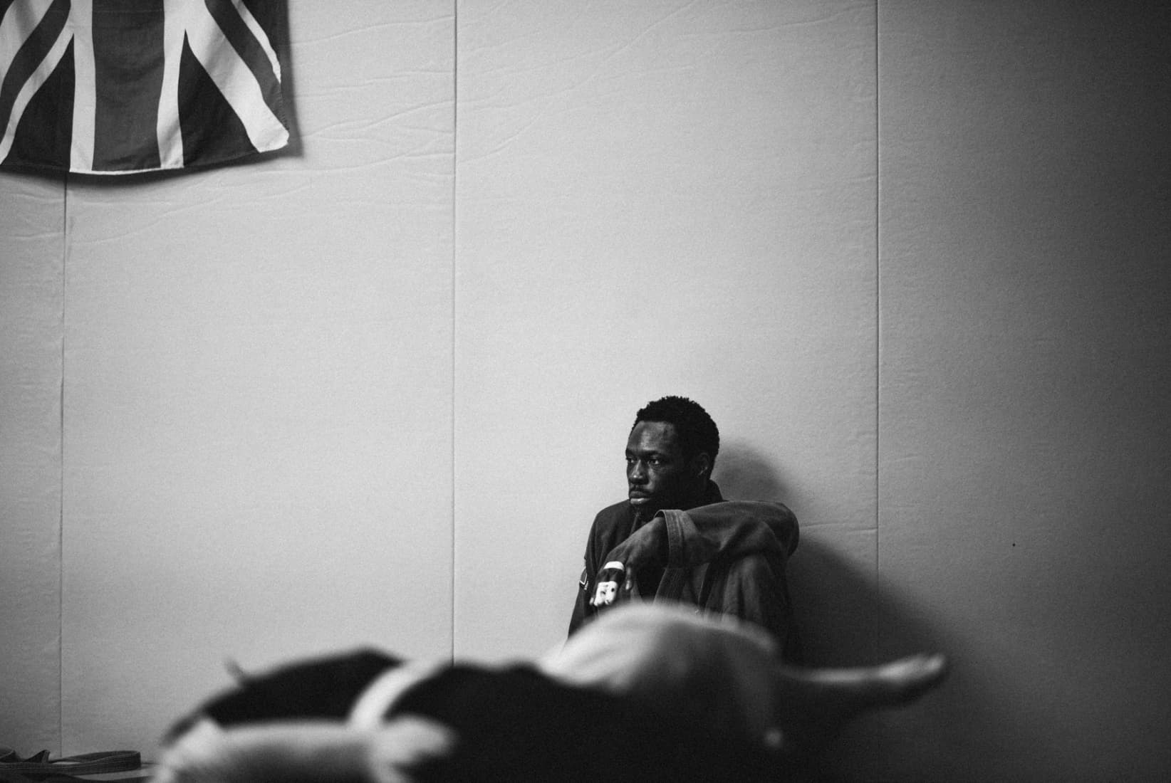 Black-and-white image of a man sitting quietly against a padded gym wall beneath a Union Jack flag, gear blurred in the foreground