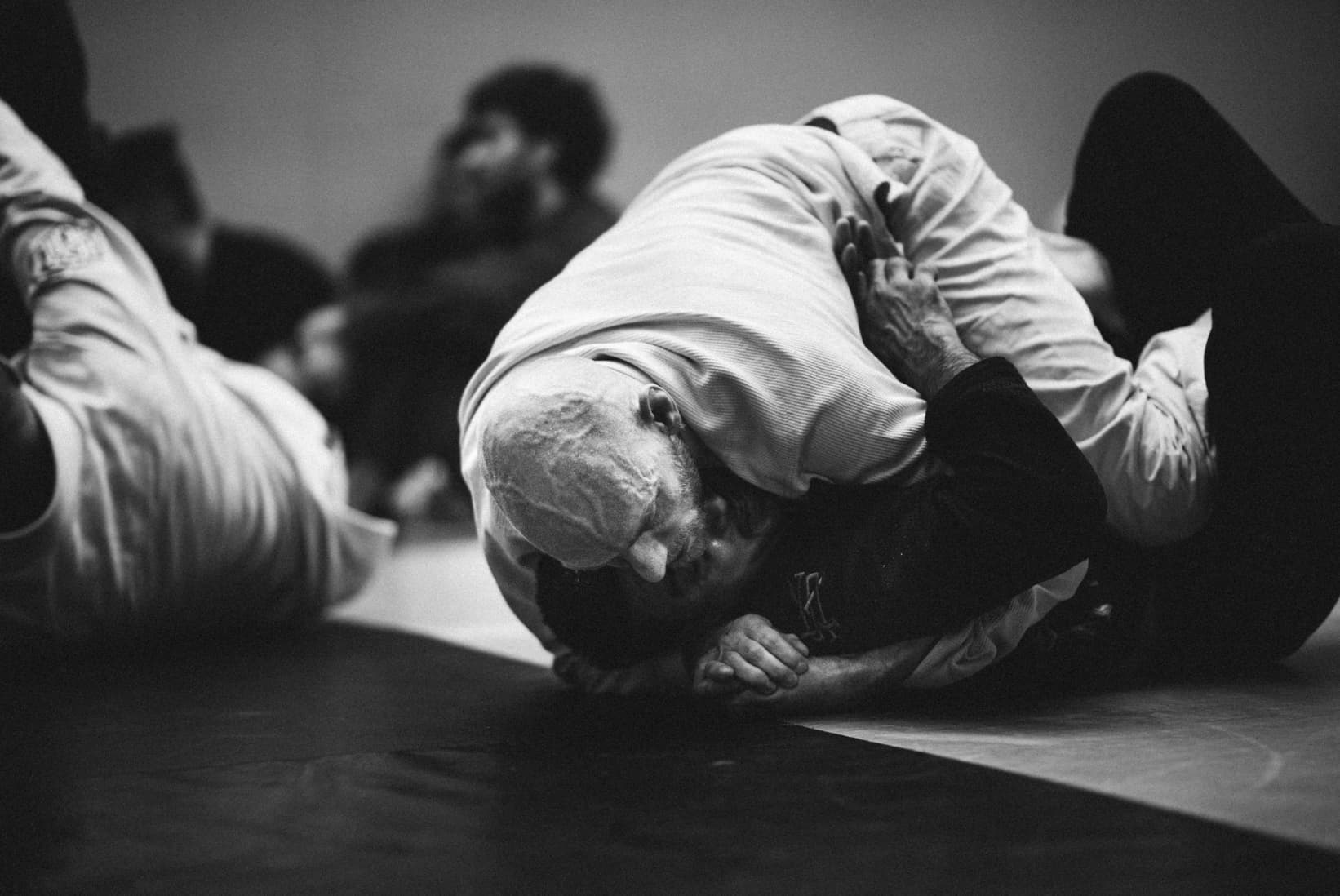 Black-and-white scramble on the mat, a bald athlete in a white gi head-to-head with an opponent in a dark gi, hands locked, other pairs rolling behind
