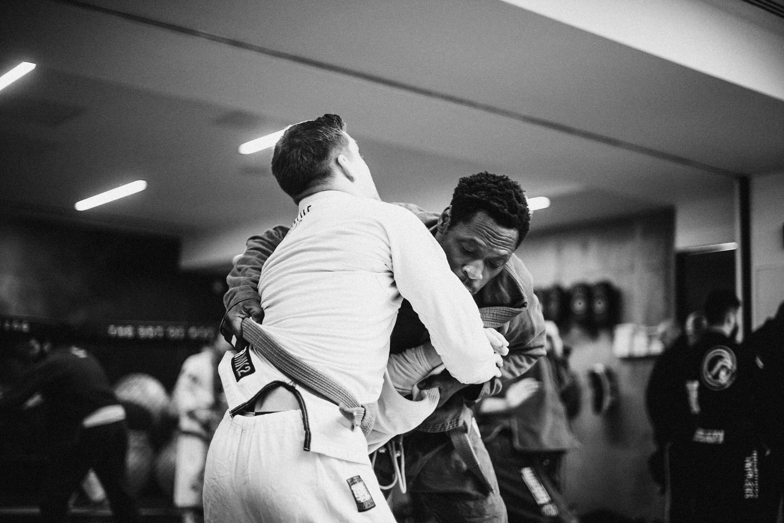 Black-and-white grip-fighting exchange between two standing athletes, one in a white gi with a grey belt turning away from his partner's collar grip