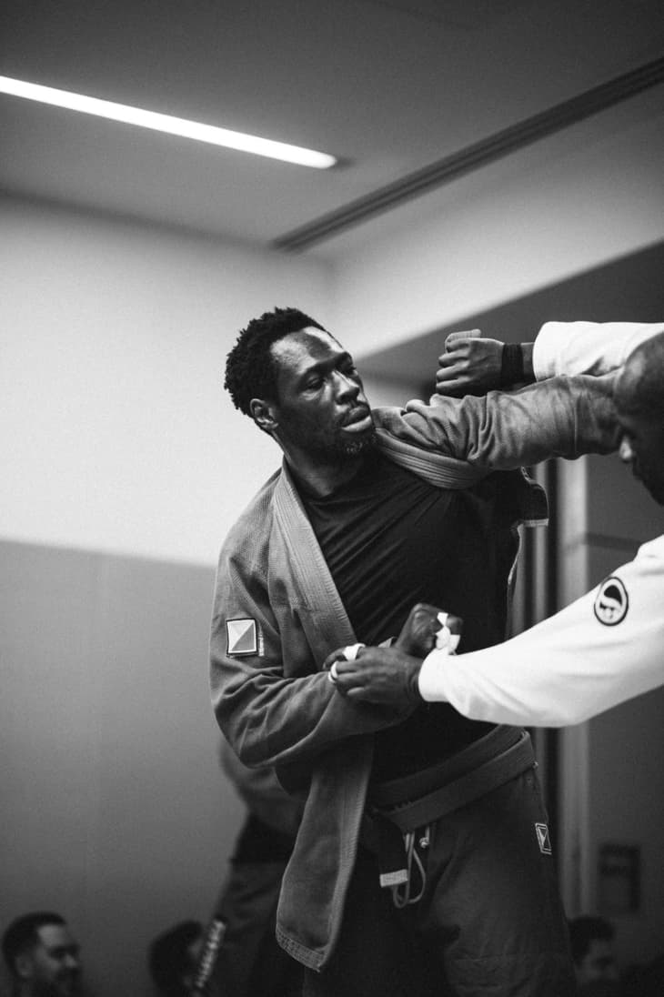 Black-and-white image of an athlete in a grey gi gripped at the collar and wrist by an opponent in a white gi, eyes fixed downward