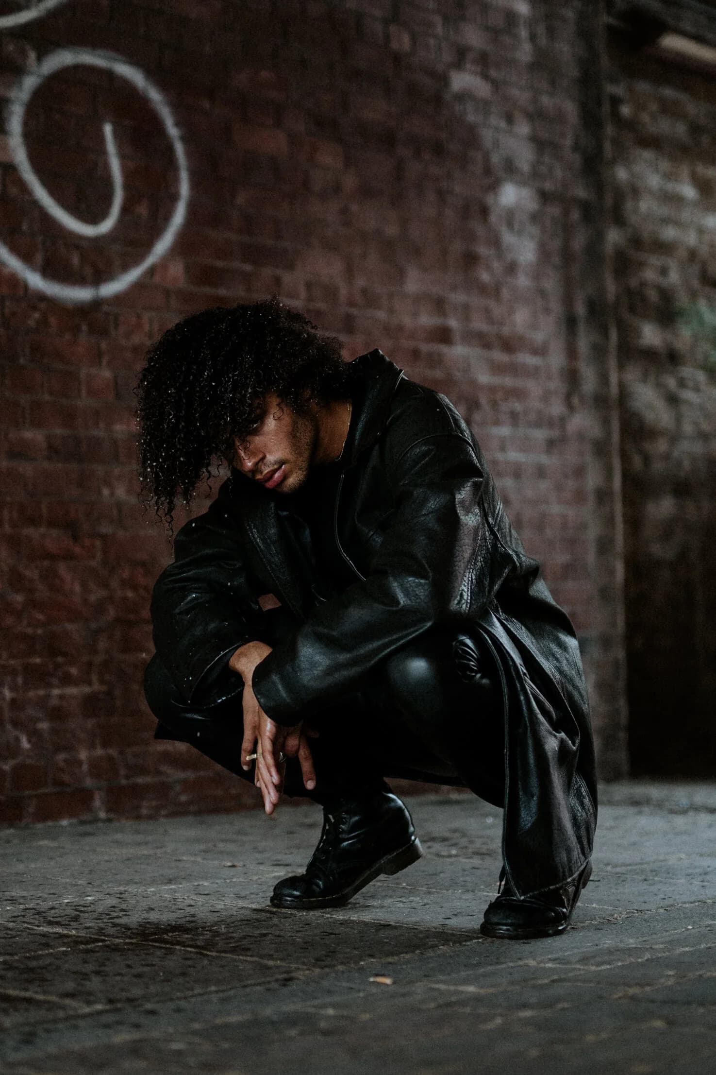 Model with curly hair crouching low against a graffitied brick wall in an oversized black leather coat and polished boots