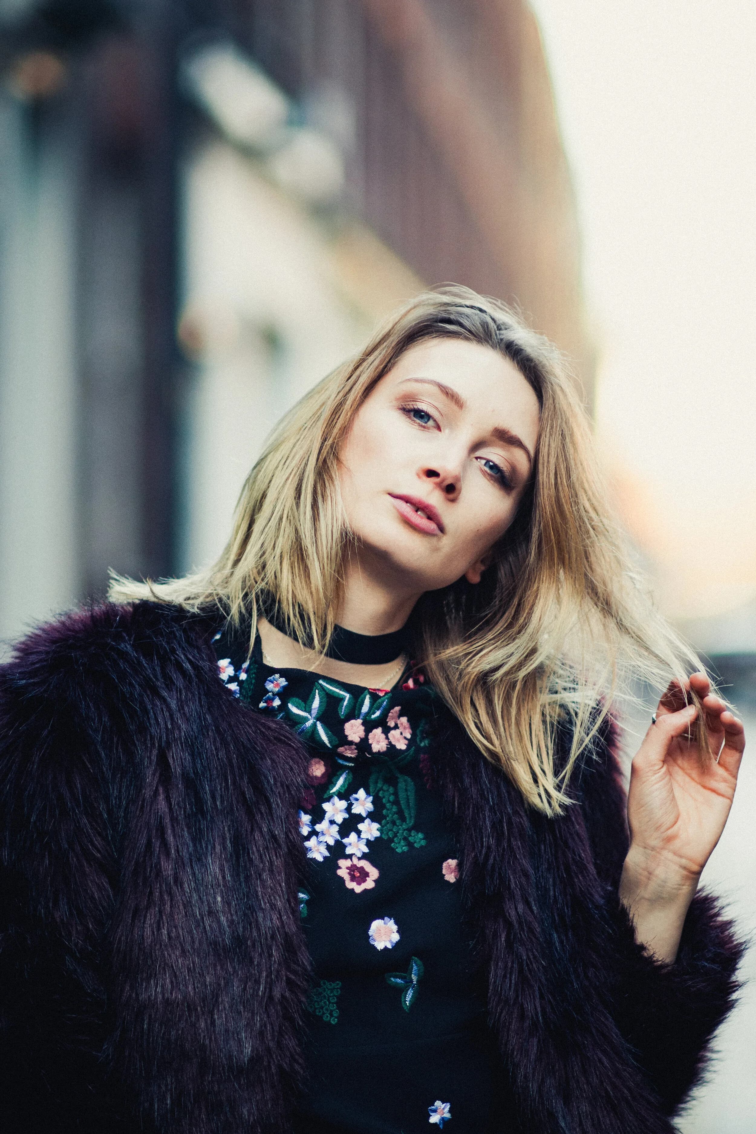 Blonde model in a plum faux-fur coat over a floral-embroidered top and black choker, tilting her head on a city street