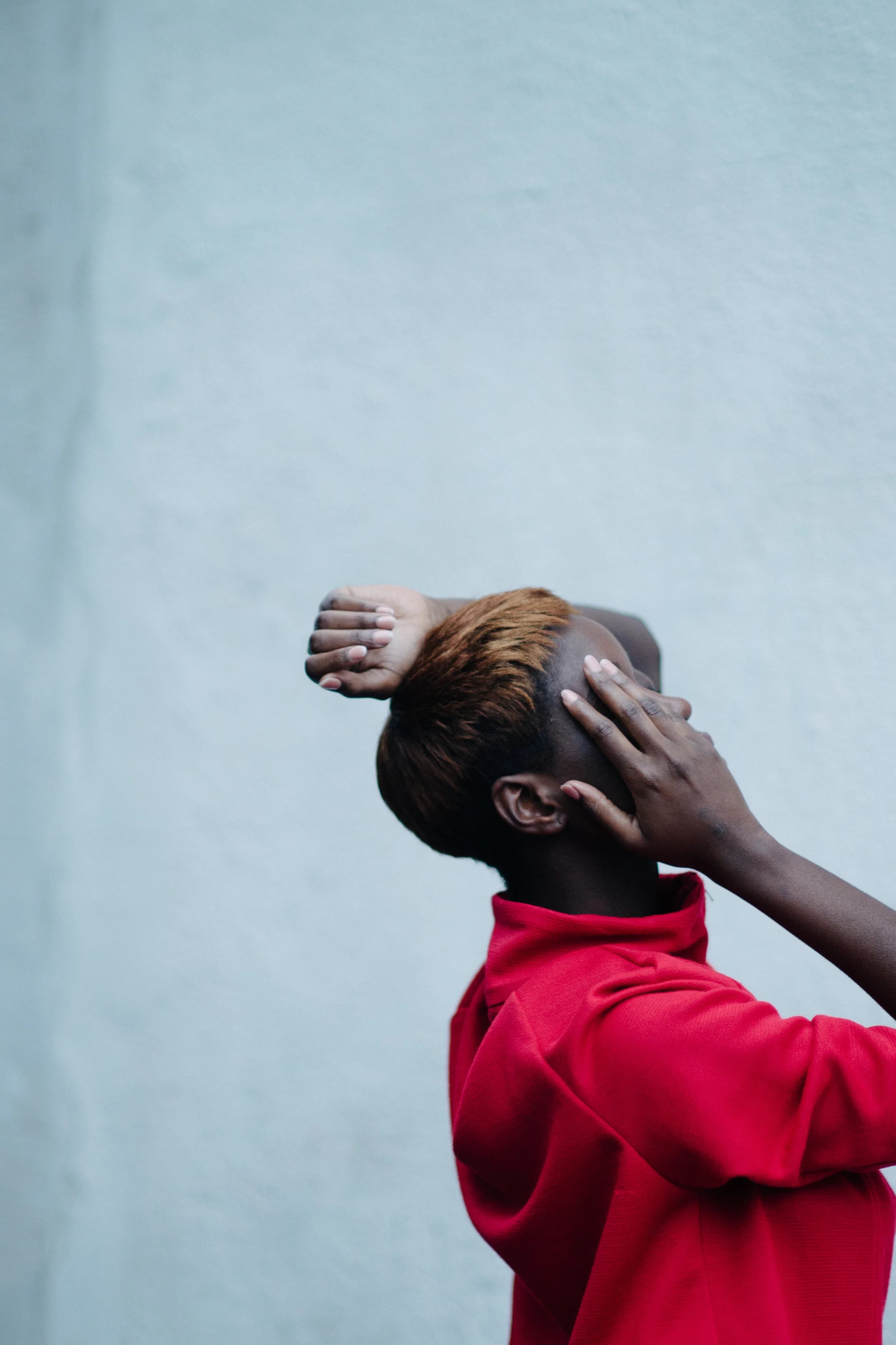 Model in a red high-collared top turning away against a pale blue wall, hands pressed to her cropped copper hair