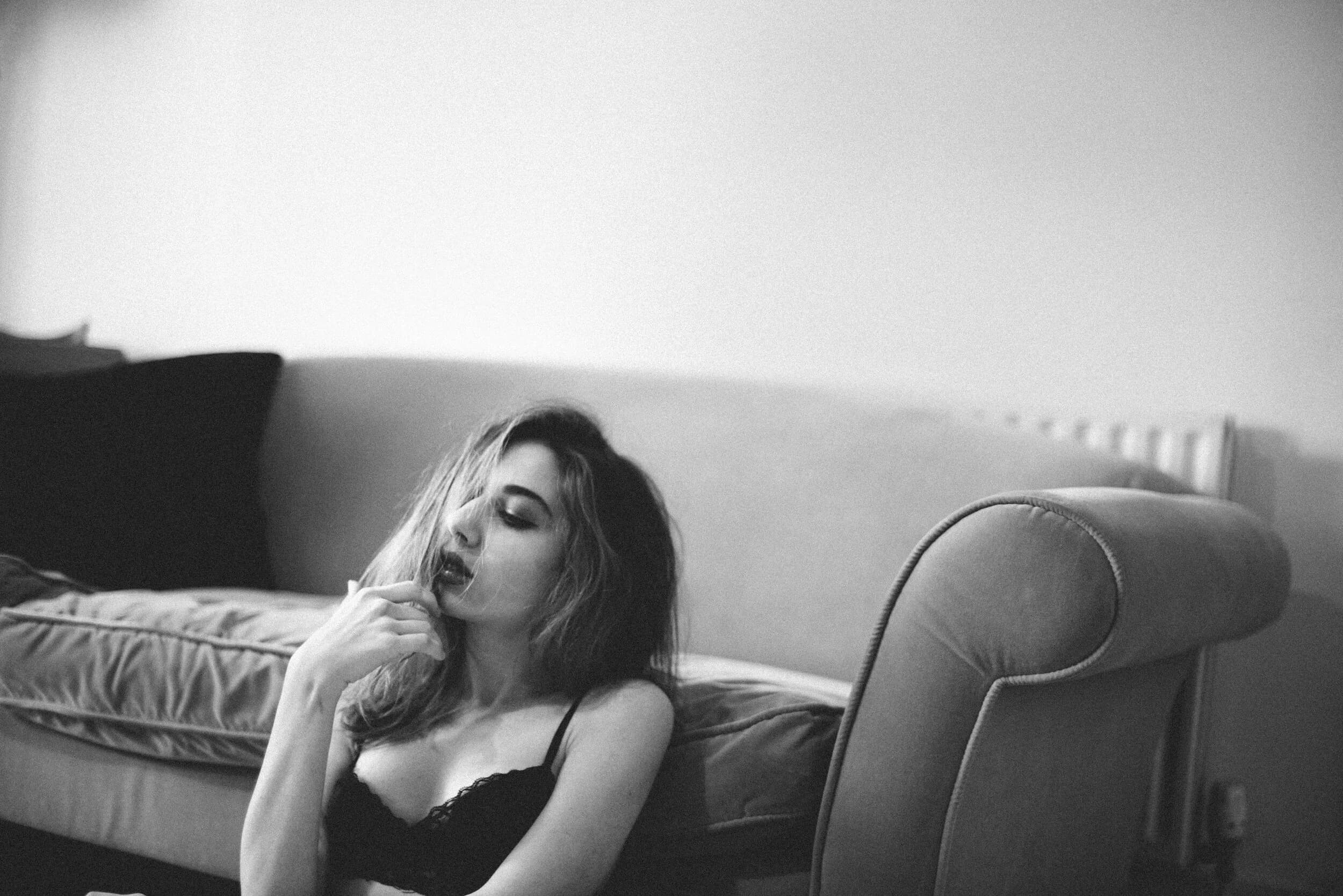 Black-and-white image of a model in a lace bralette sitting on the floor against a velvet sofa, hair falling across her face