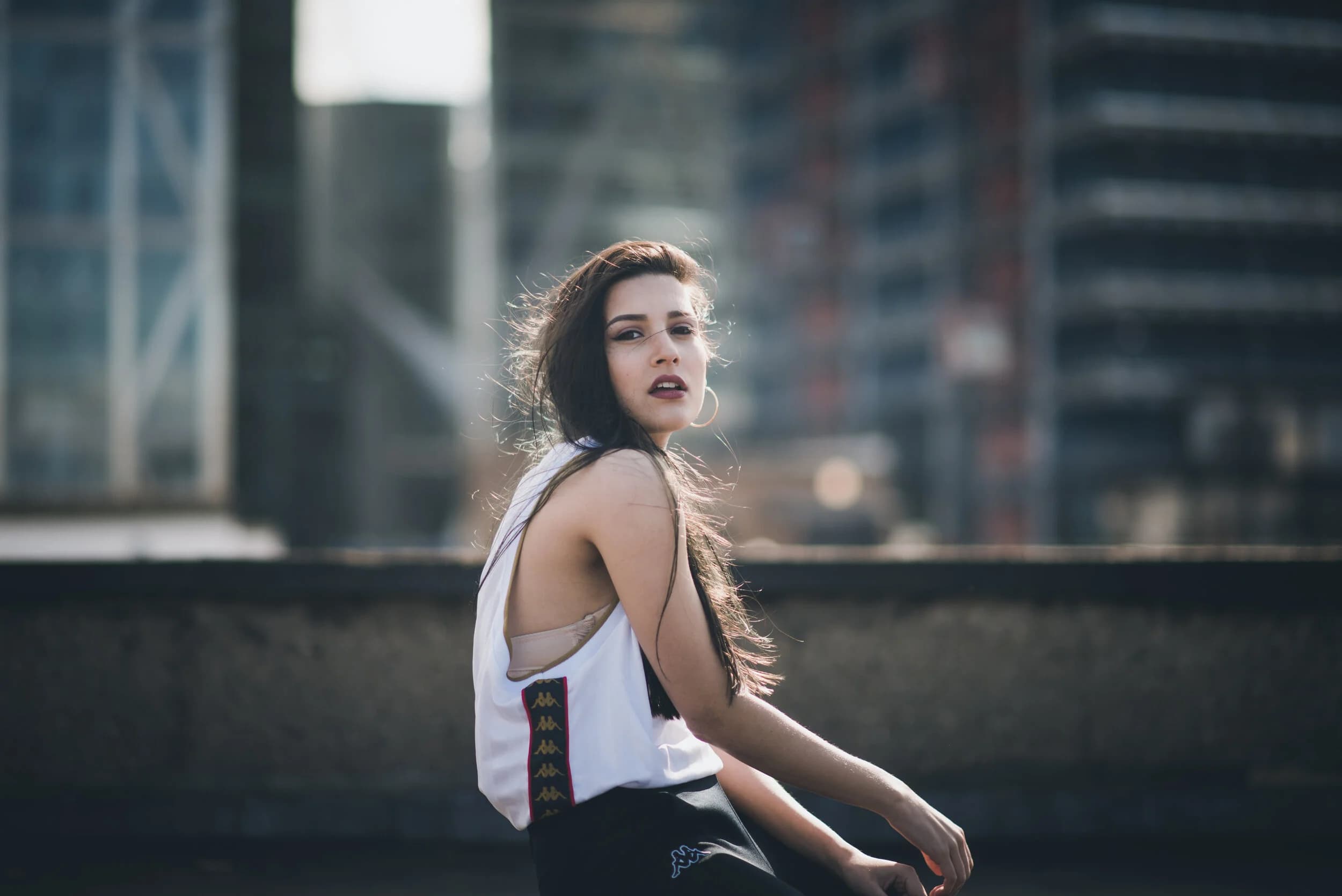 Model in a white Kappa vest and hoop earrings glancing over her shoulder, hair blowing across glass towers behind