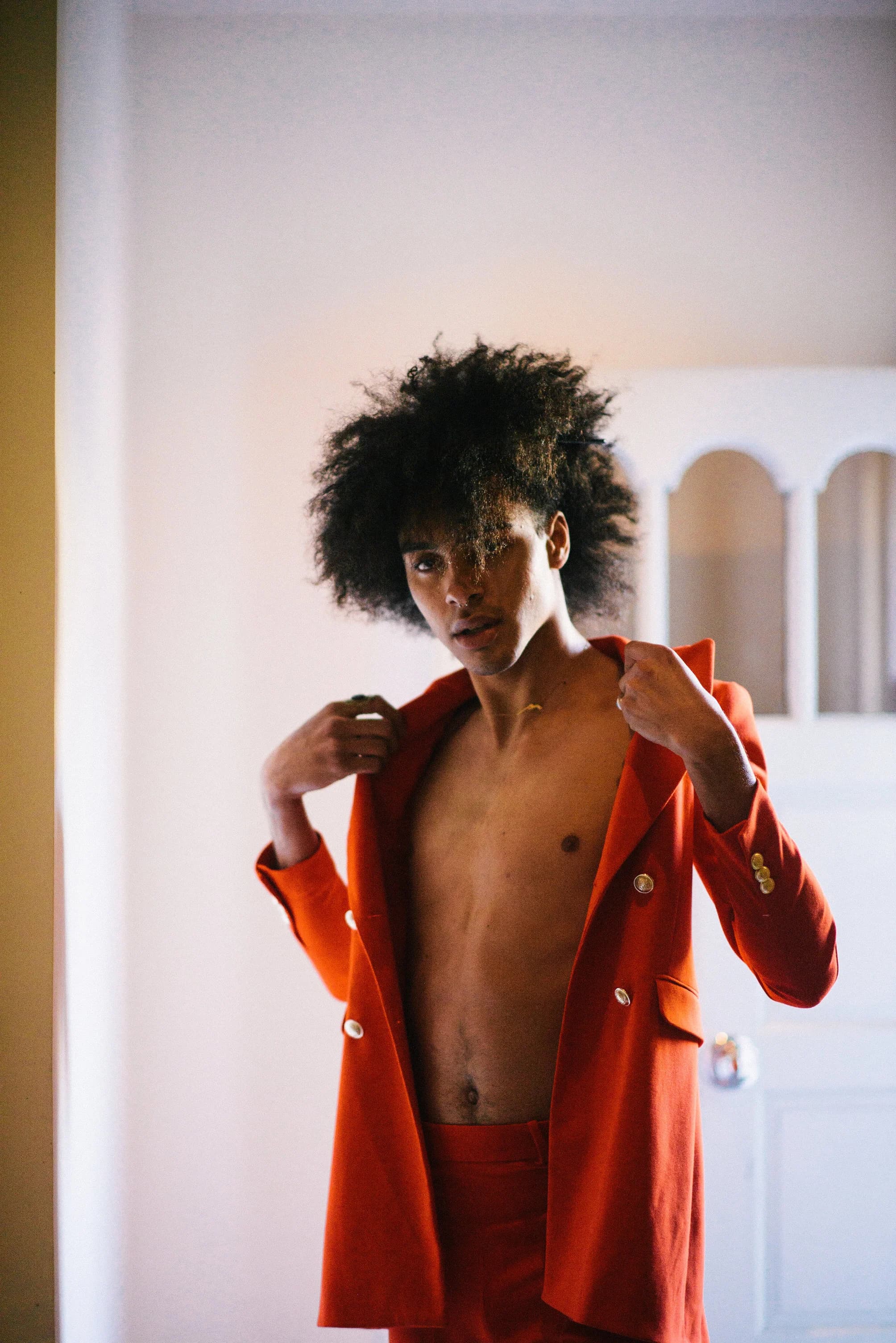 Model with a full afro pulling a rust-red double-breasted blazer over his bare chest in front of a white arched door