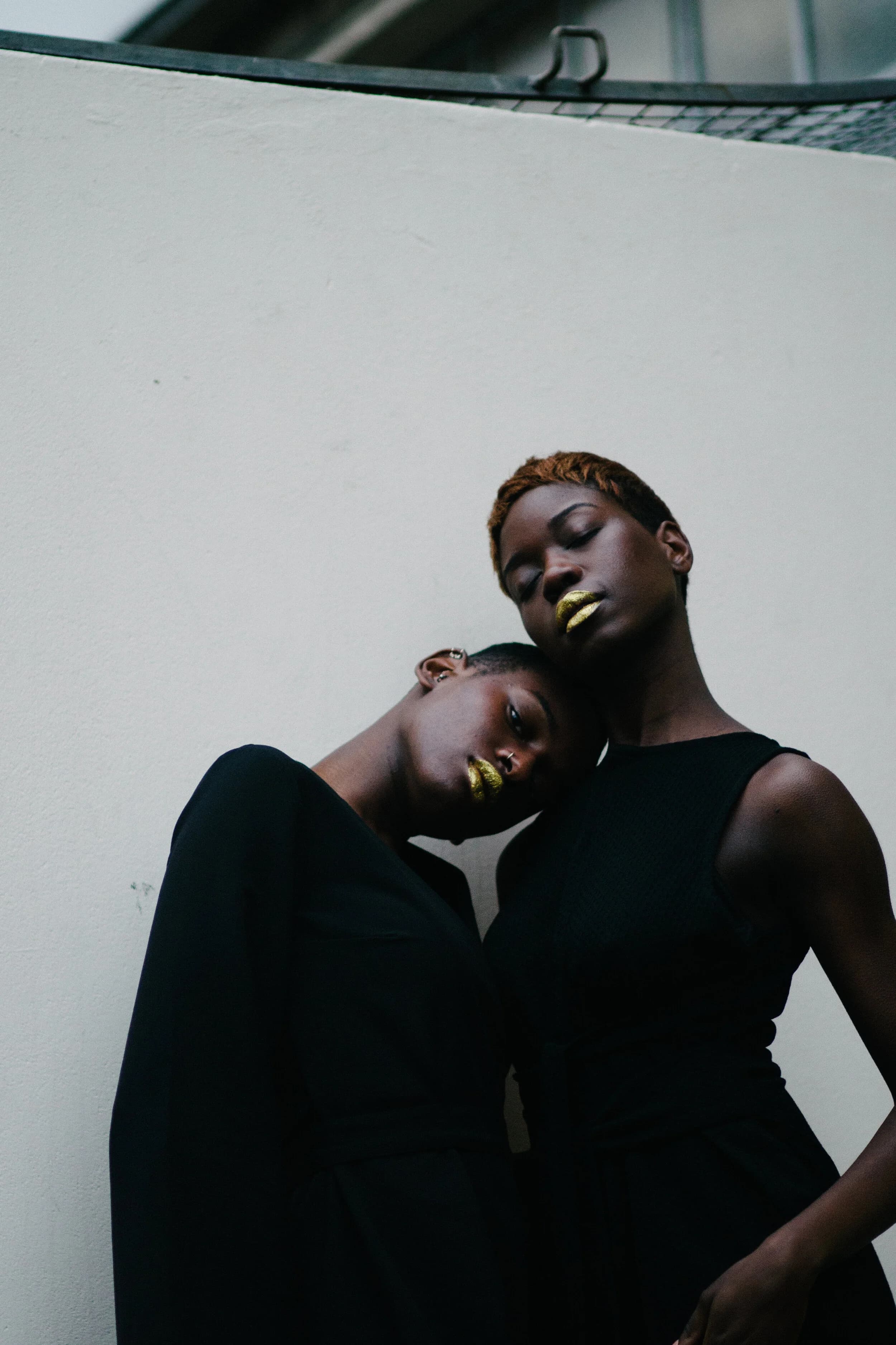 Two models in black with gold-painted lips, one resting her head on the other's shoulder against a white courtyard wall