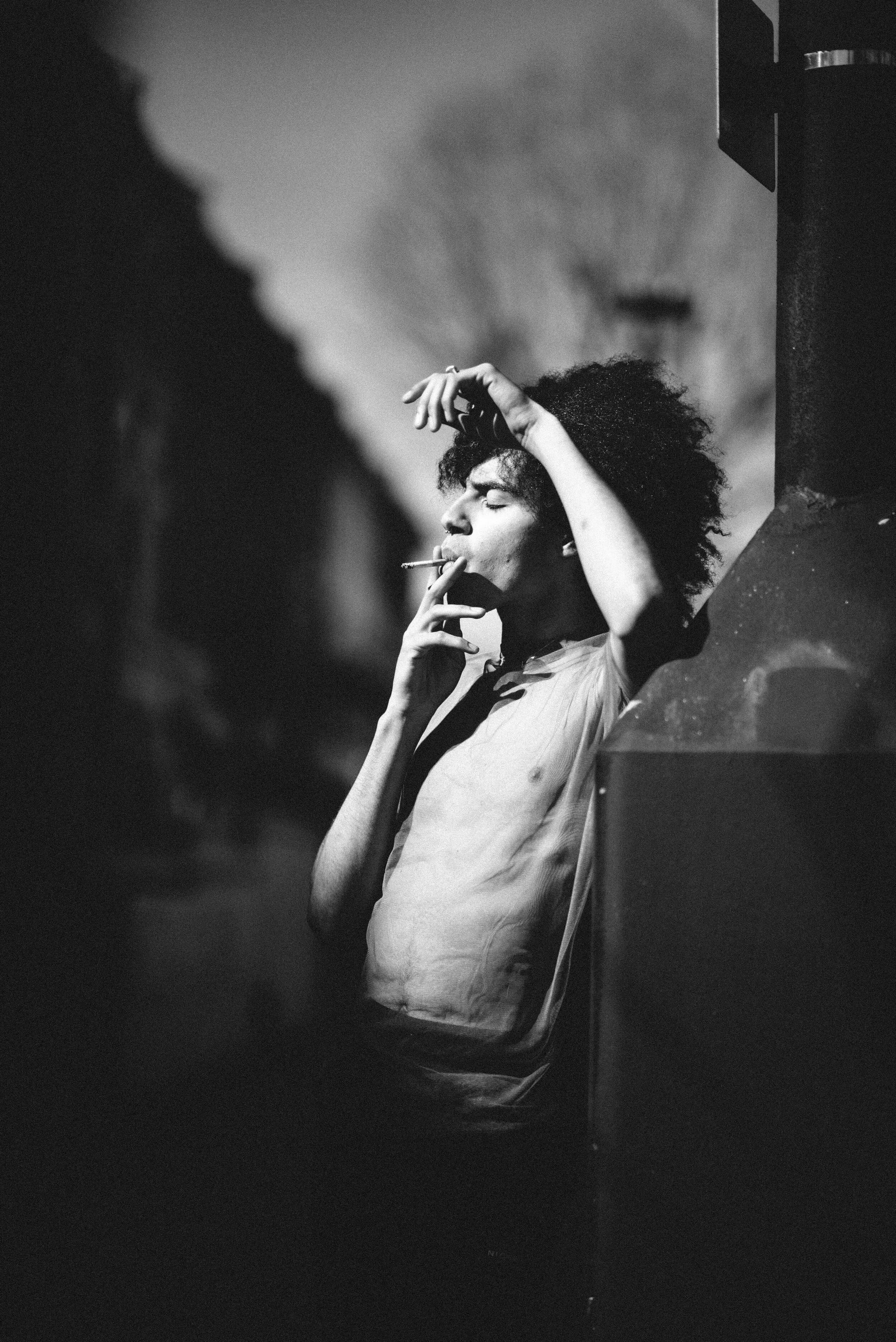 Black-and-white profile of a model in a sheer shirt leaning against a dark column, smoking with one arm raised overhead