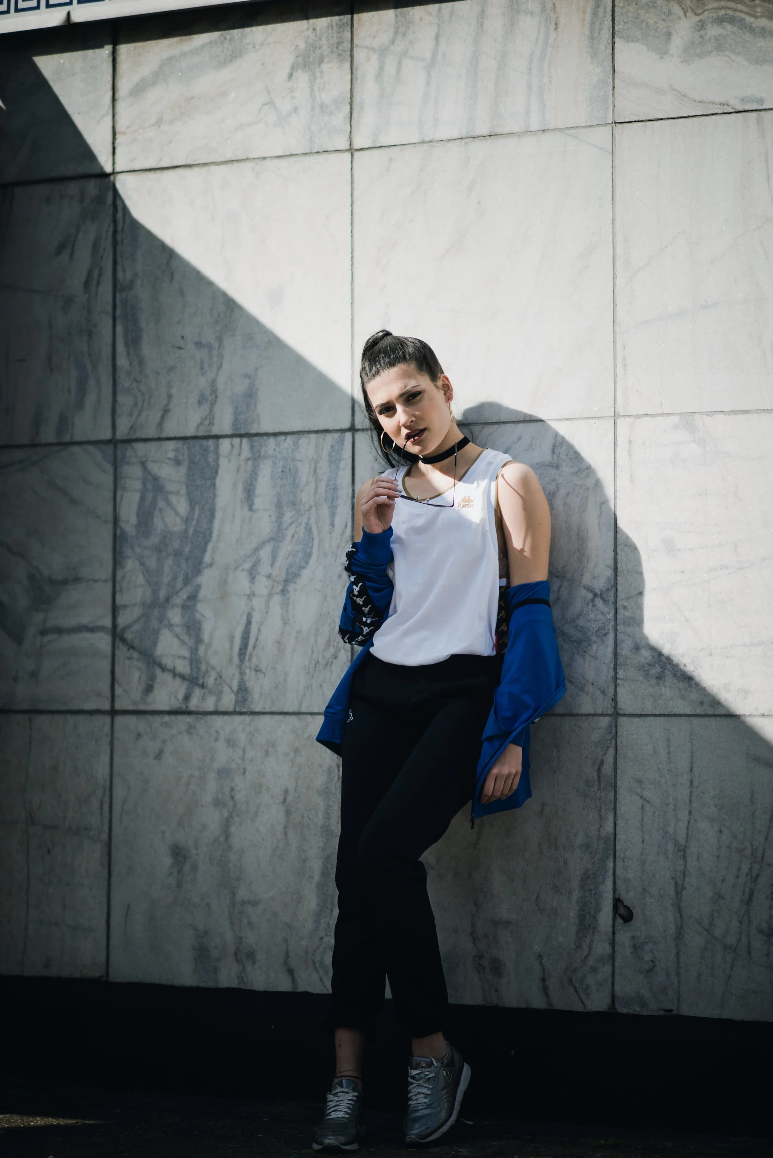 Model in a white Kappa tank, black choker and blue track jacket pulled off her shoulders, leaning on a marble wall cut by a diagonal shadow