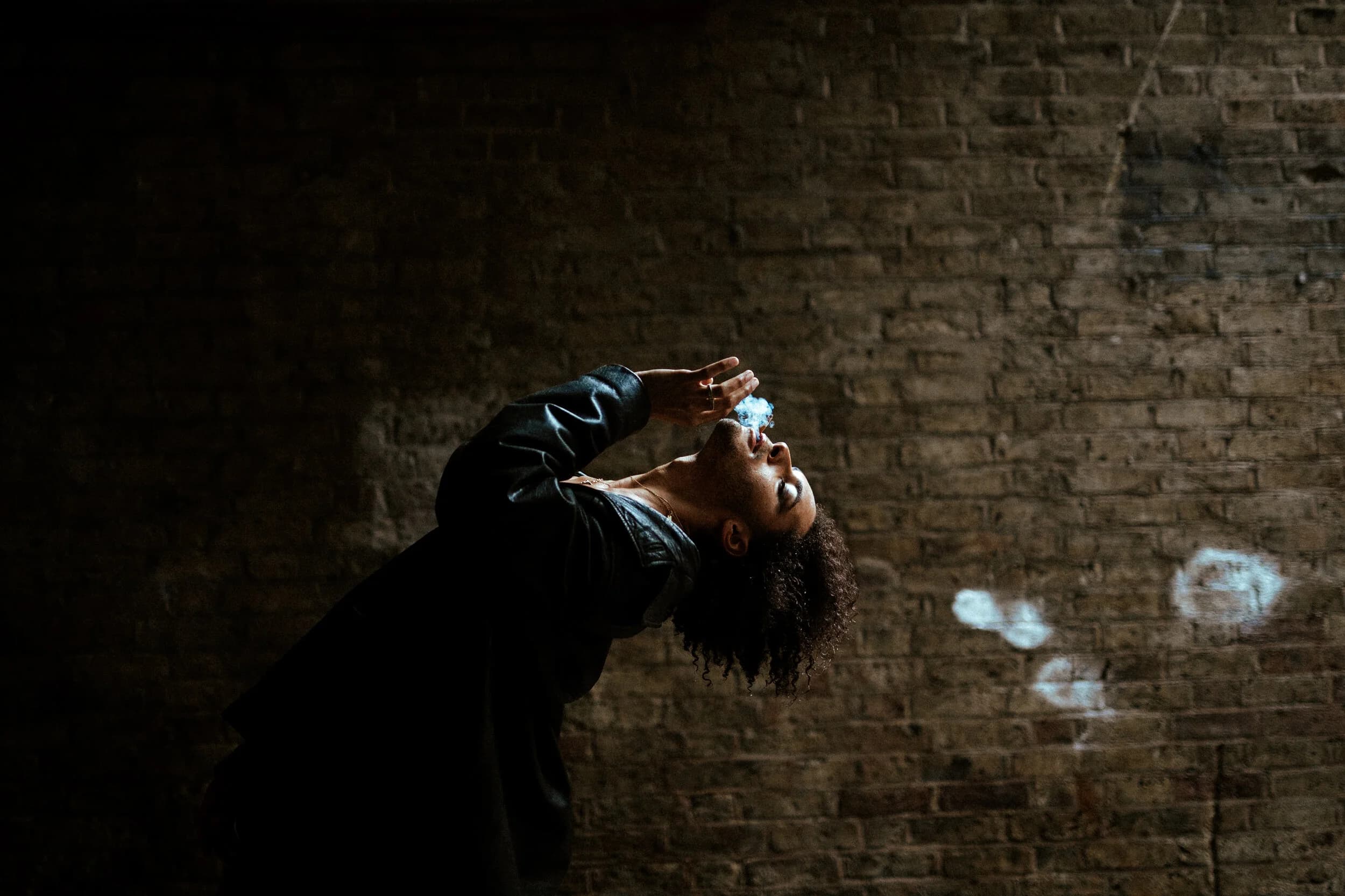Model in a leather jacket bending backwards against a dark brick wall, exhaling smoke toward the light