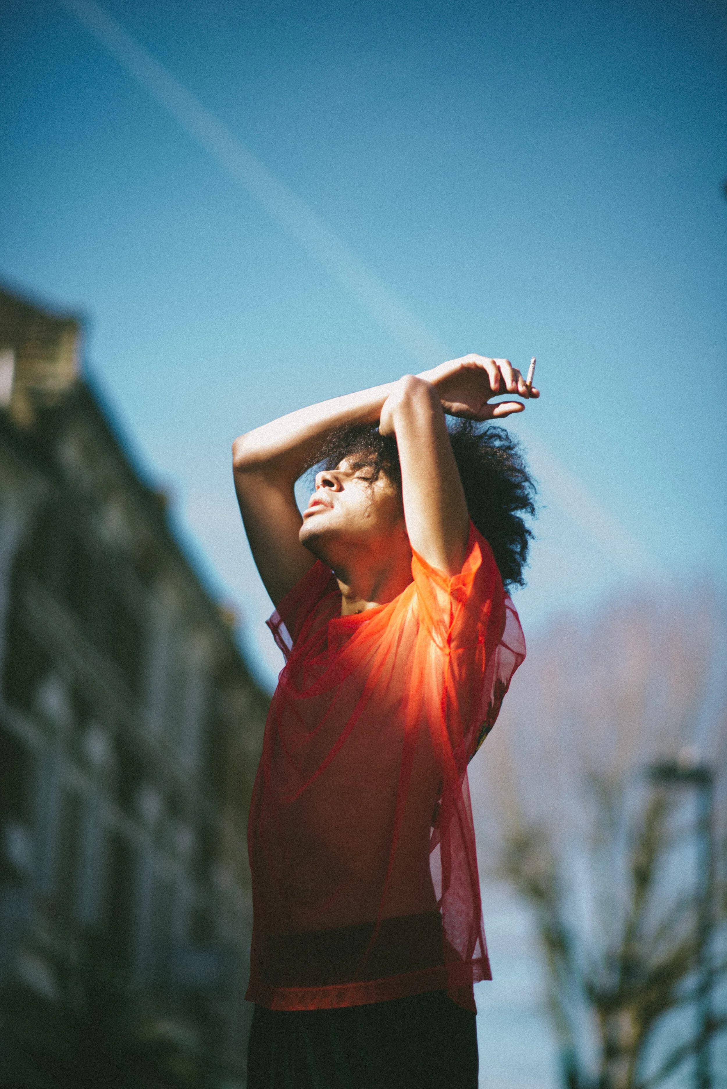 Model in a sheer red top with eyes closed against a blue sky, arms crossed above his head holding a cigarette