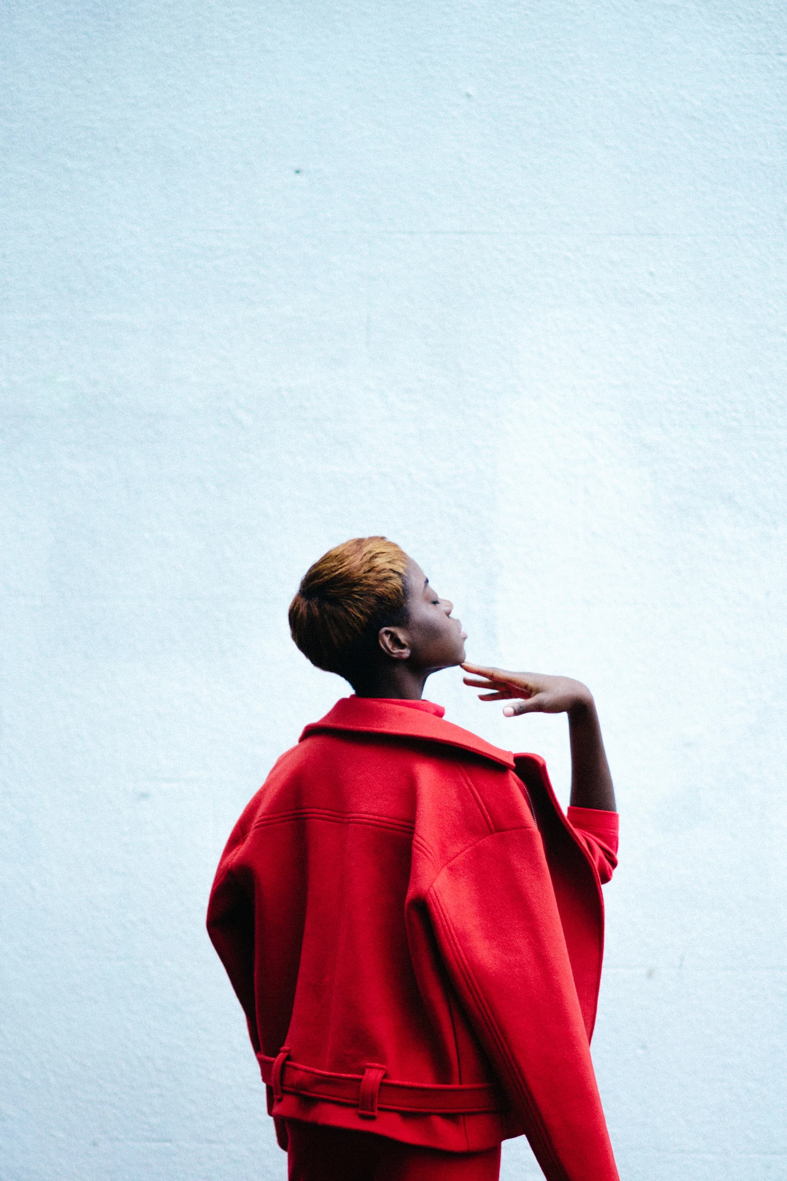 Model in an oversized belted red jacket seen from behind against a white wall, chin lifted with a hand raised beneath it