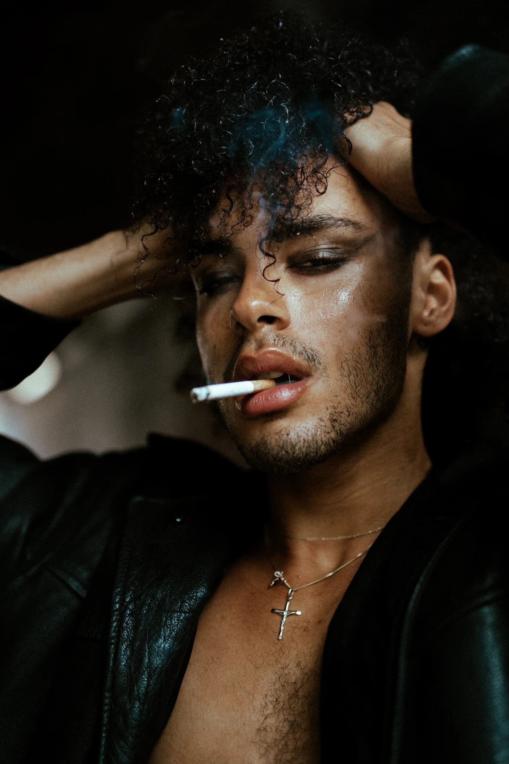 Close portrait of a model with smoky eye makeup and a cigarette between his lips, hands running through his curly hair, a crucifix at his open leather jacket