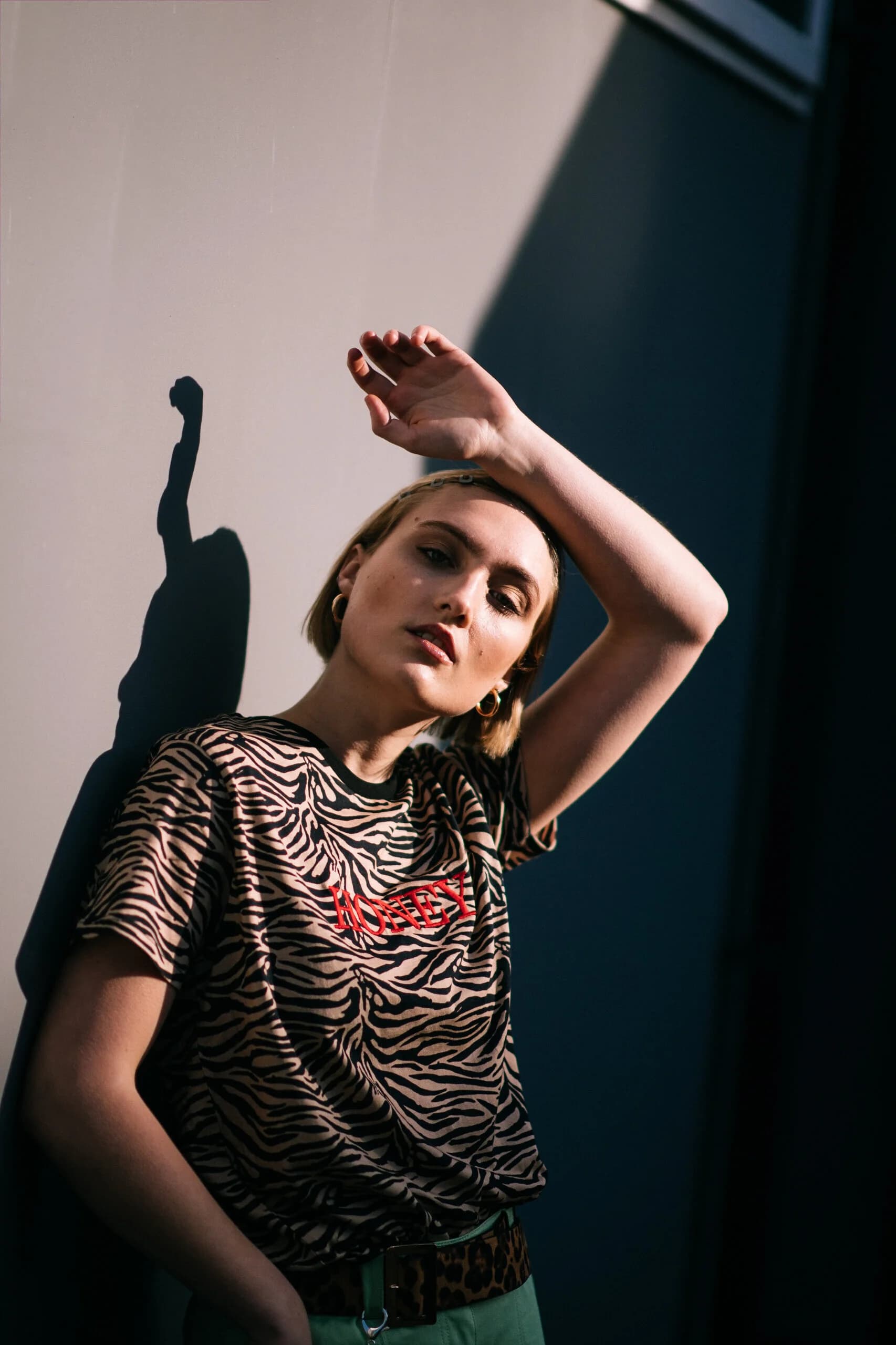 Model in a zebra-print HONEY t-shirt with an arm resting over her brow, her shadow cast sharply on the sunlit wall behind