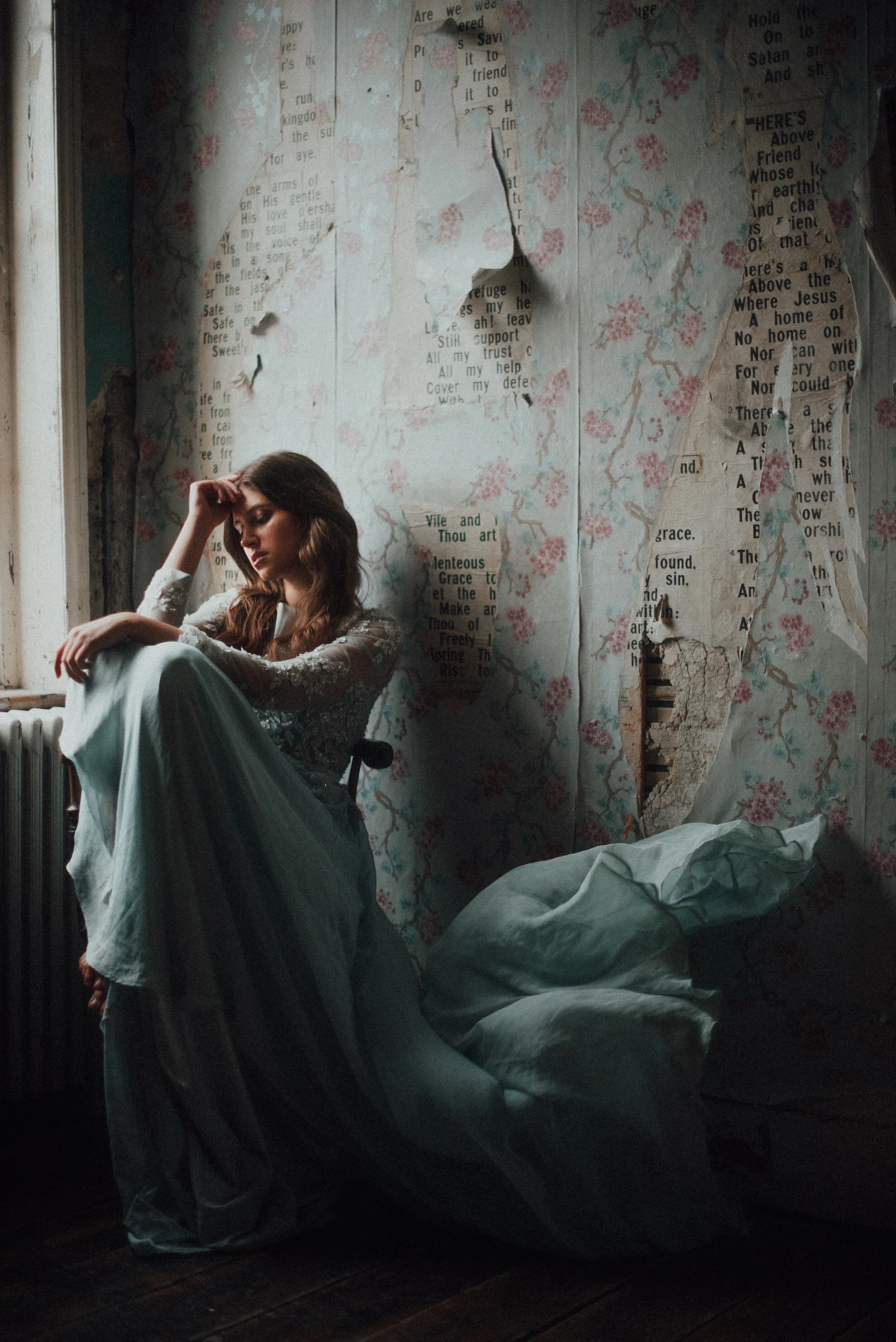 Model in a pale blue gown with an embroidered lace bodice sitting pensively by a window, torn floral wallpaper and hymn-sheet fragments covering the wall