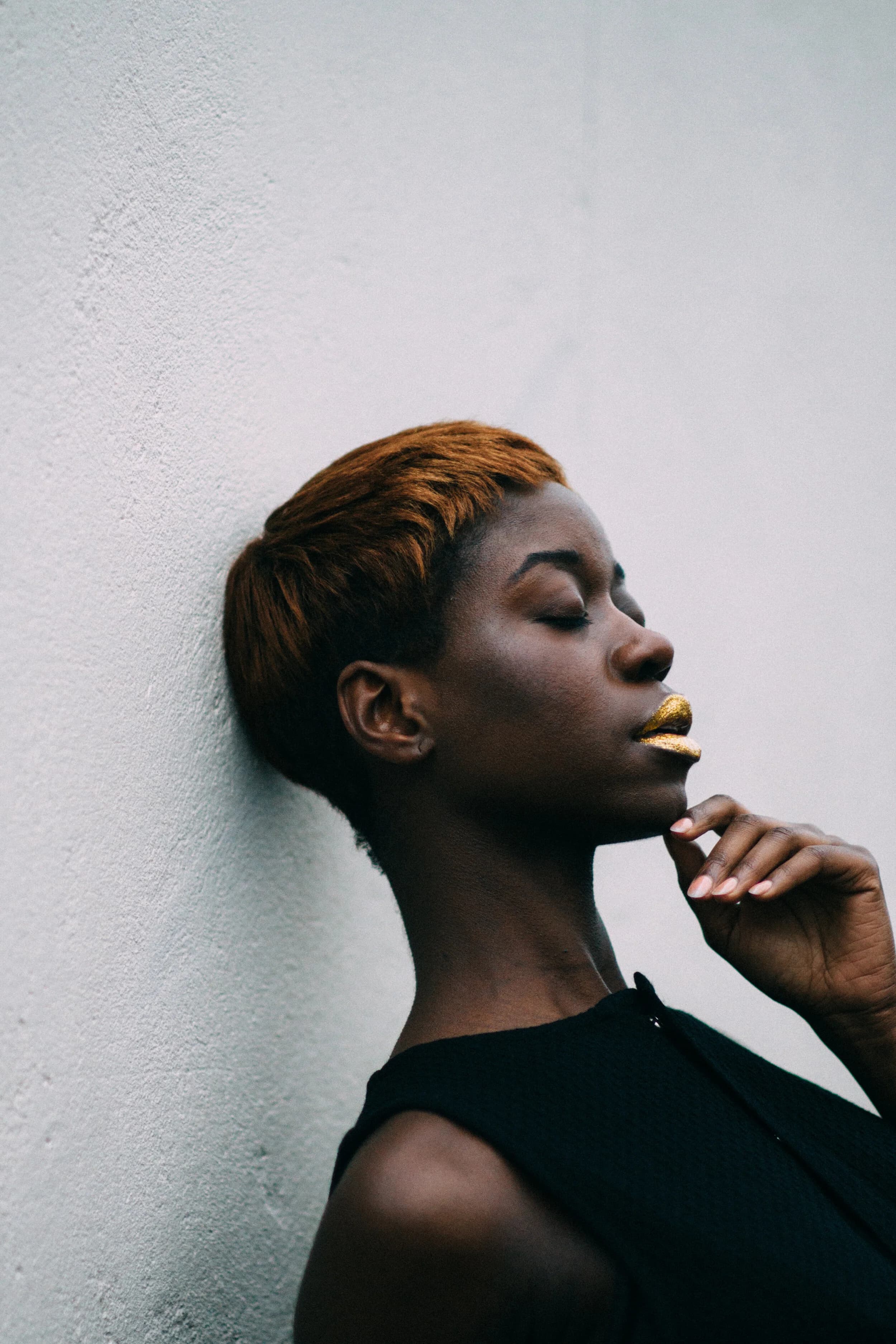 Profile portrait of a model with cropped copper hair and gold-glitter lips, eyes closed against a white wall, one hand raised to her chin