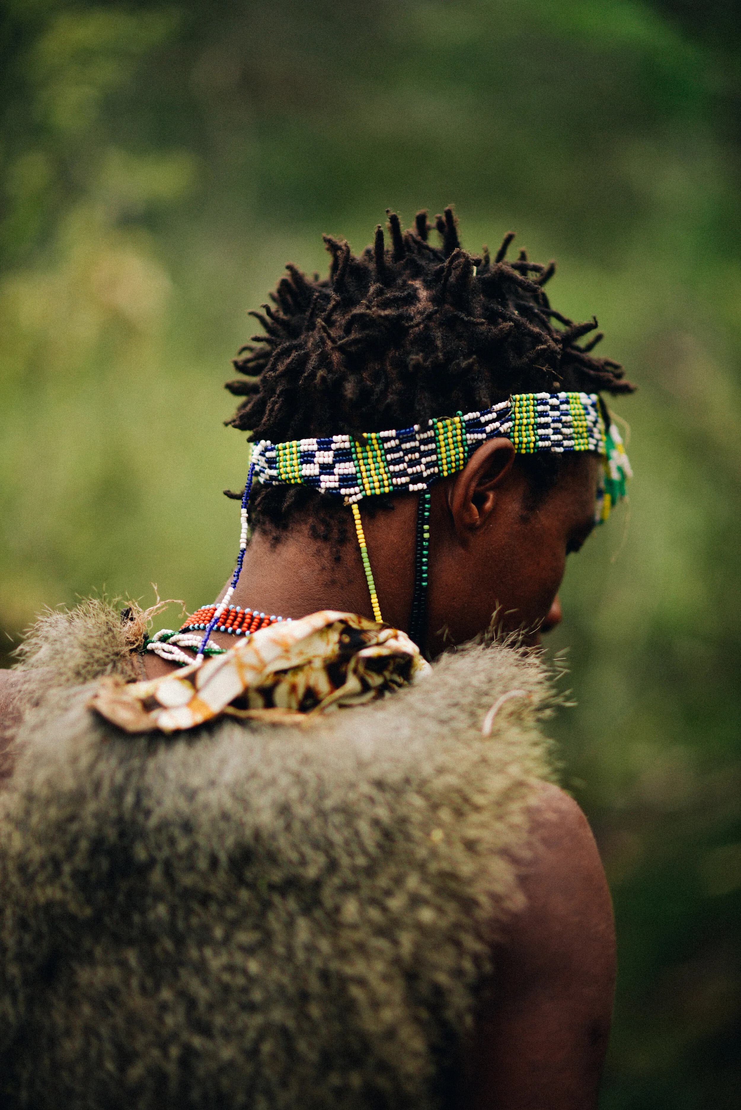 Close view from behind of a hunter's beaded blue, green and white headband over thick dreadlocked hair, with an orange bead necklace and a grey fur cape over his shoulder