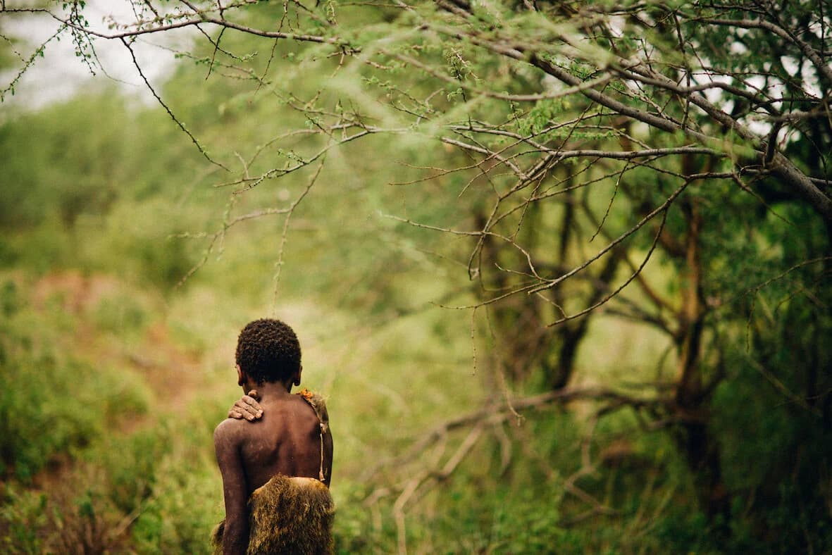 Young boy walking away beneath bare acacia branches, one hand resting on his own shoulder and a fur garment hanging at his back