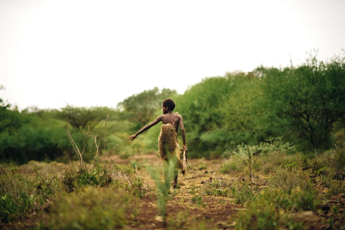 Barefoot boy in a fur wrap walking down a dirt track through open bushland, one arm swung out mid-stride