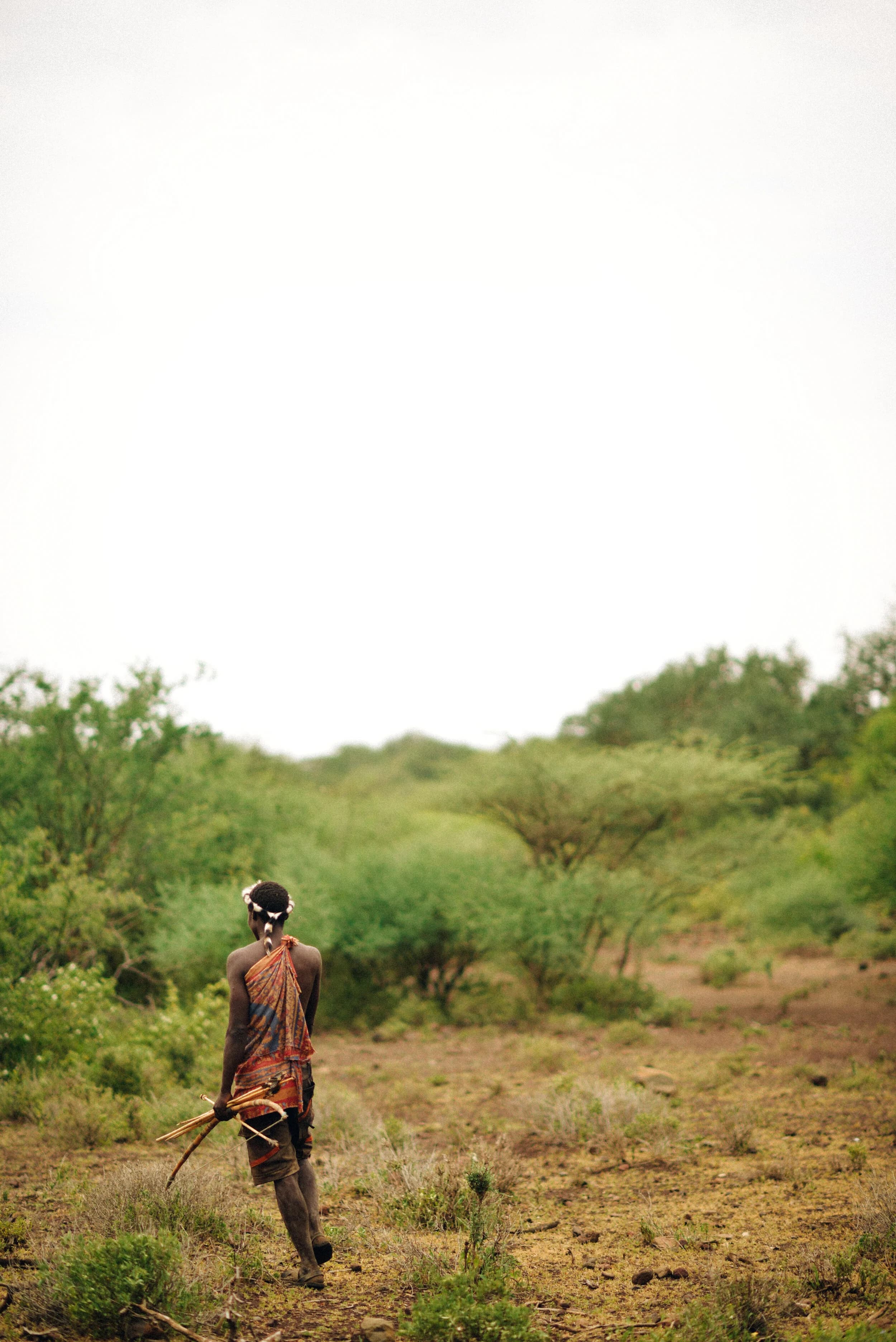 Hunter walking away into the bush carrying his bow and arrows at his side, wearing a black-and-white striped headband with a hanging braid and a patterned orange wrap