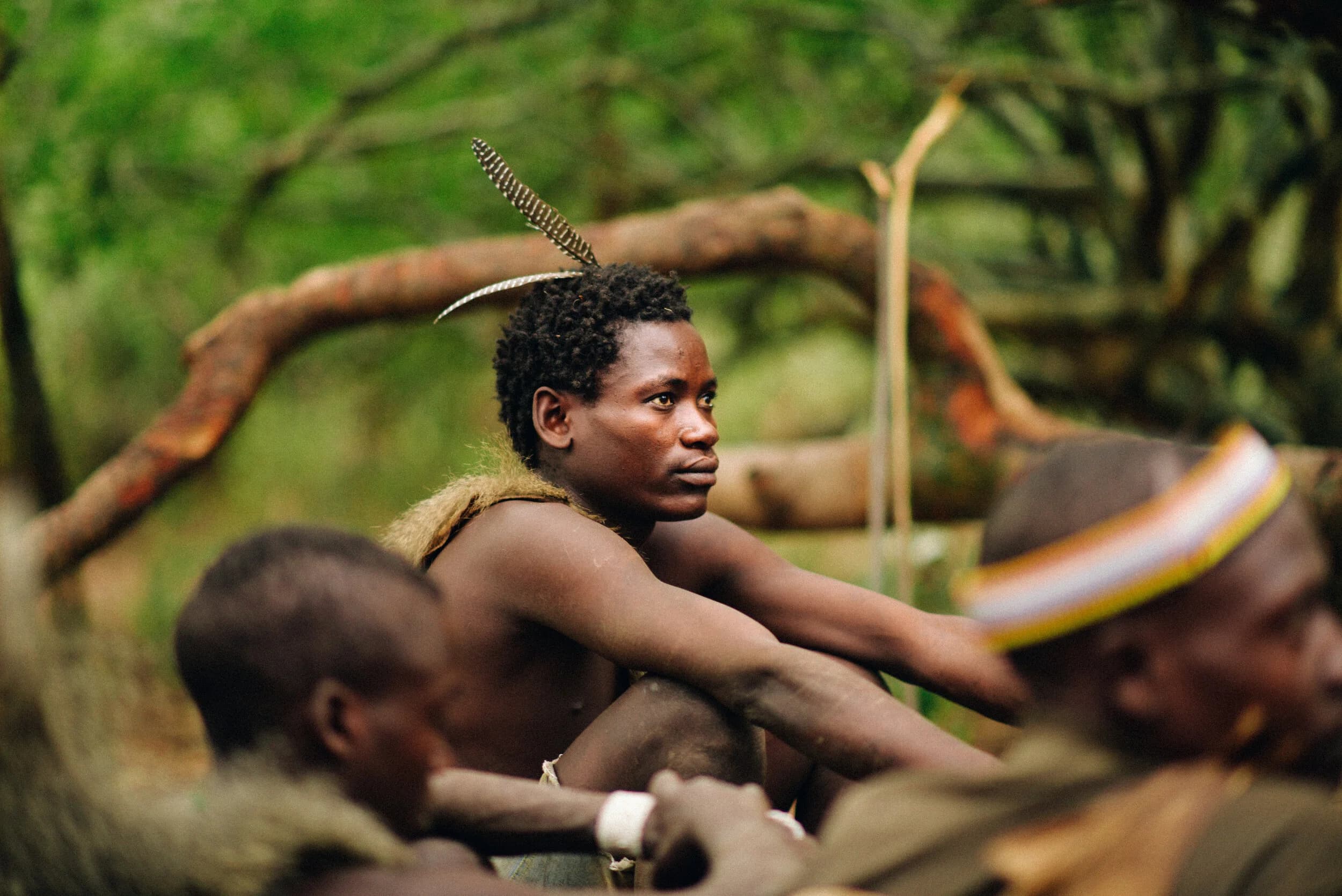 Young hunter with a single striped feather in his hair looking off in profile, seated among other resting hunters, a beaded headband in the blurred foreground