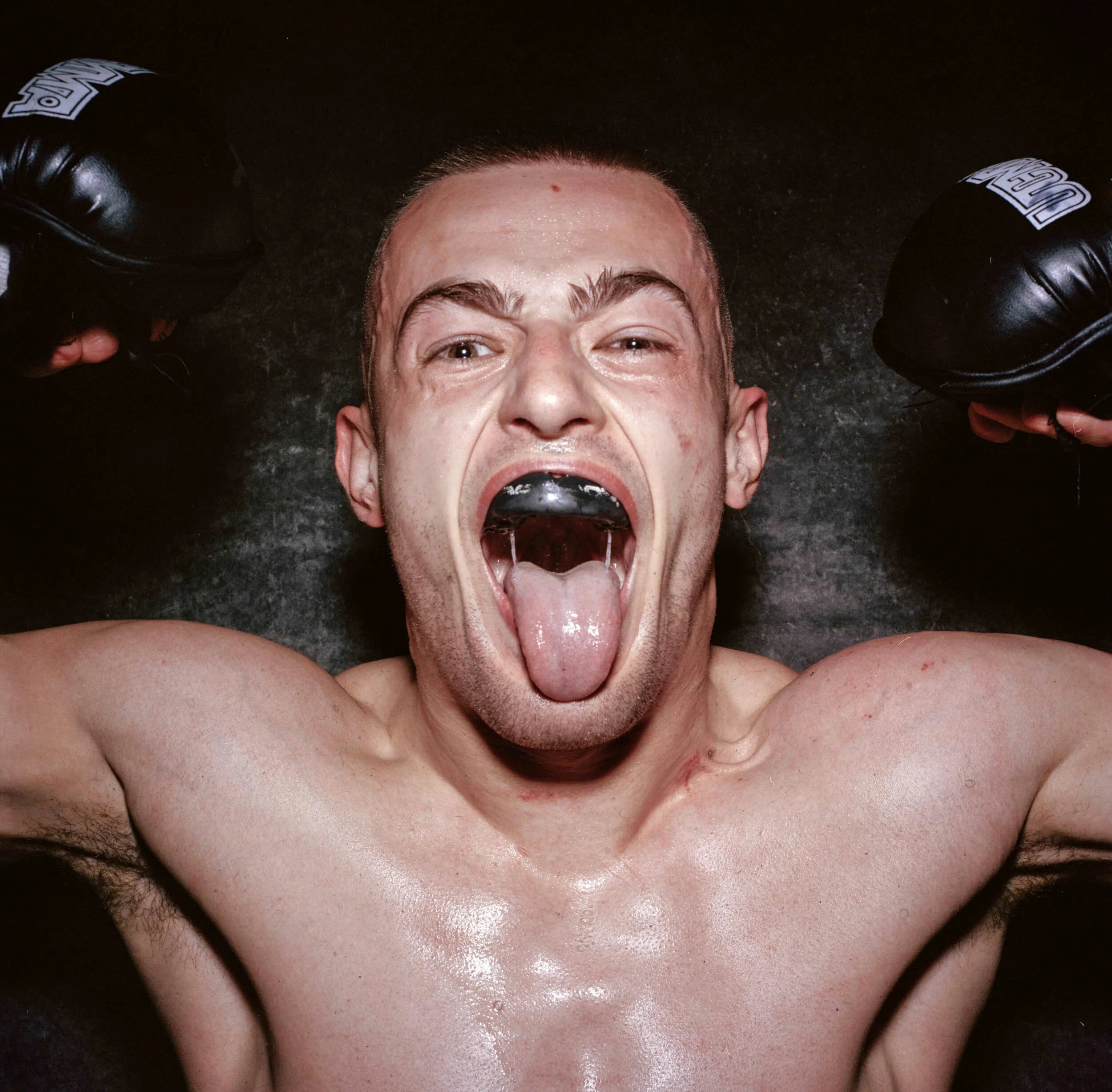 Fighter photographed from above with his tongue out over a black mouthguard, black-gloved fists raised either side of his head