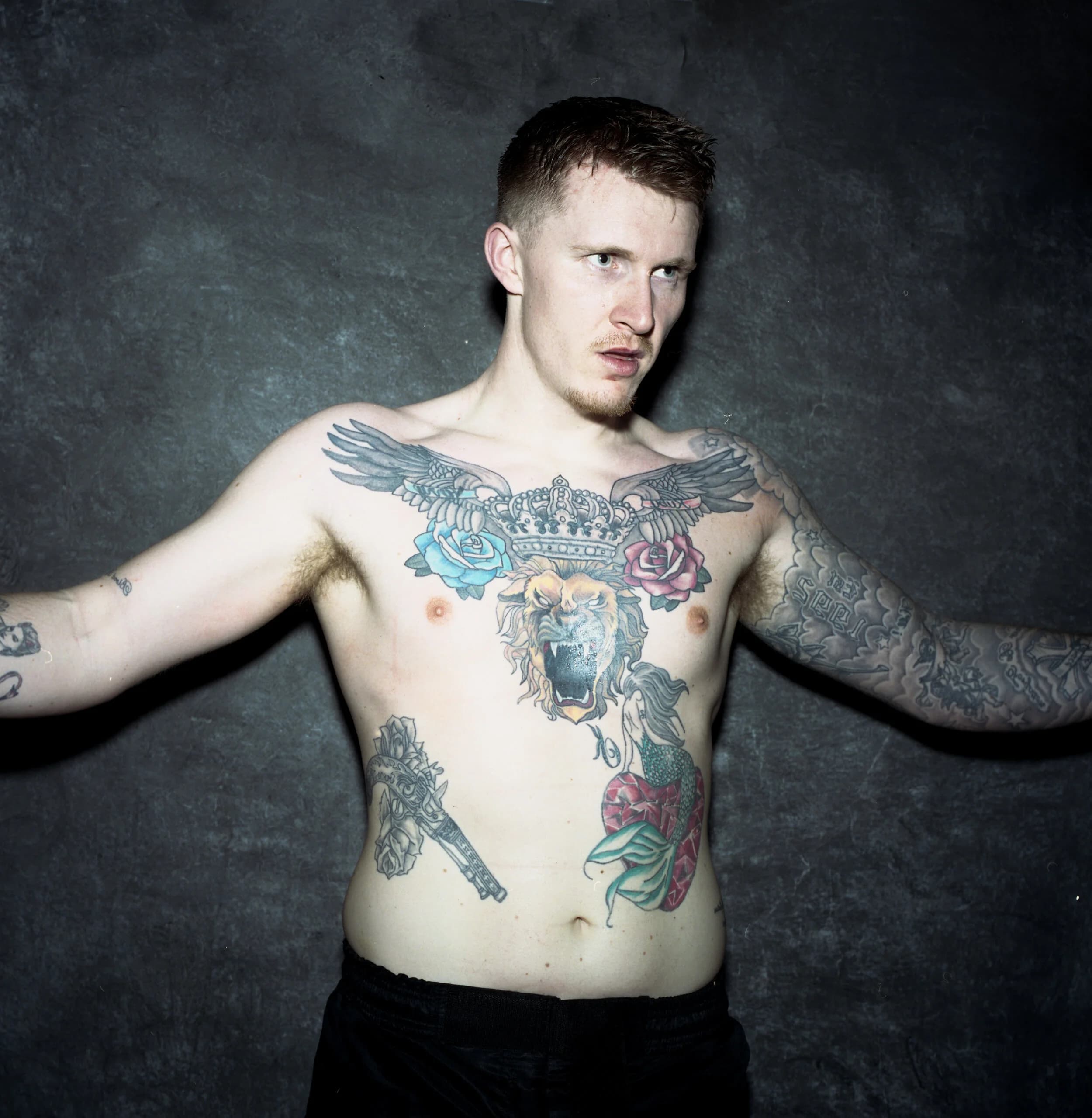 Shirtless fighter with winged-crown, lion, roses, revolver and mermaid tattoos across his chest and stomach, arms spread wide as he glances to the side