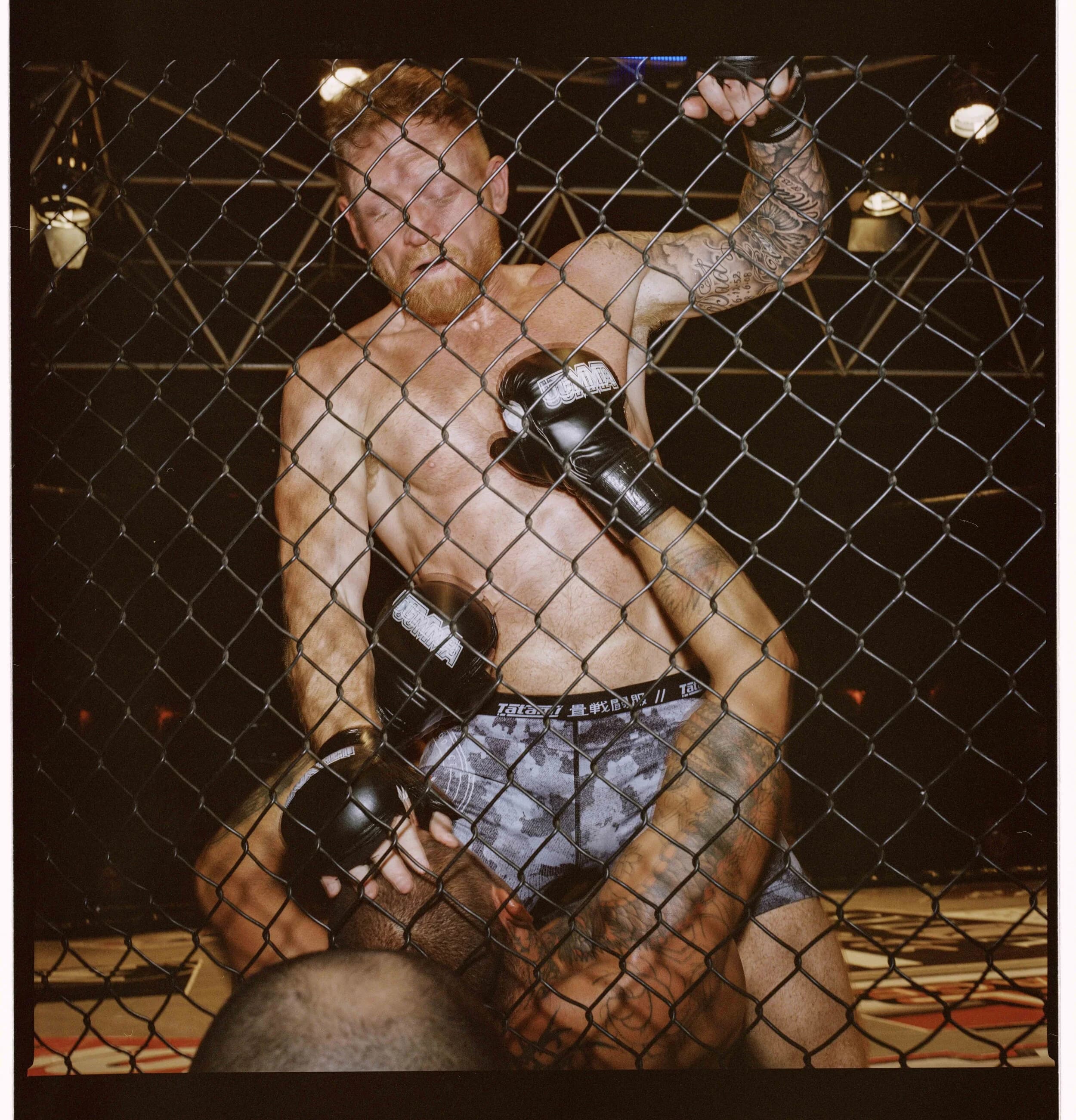 Fight seen through the chain-link cage, one fighter pressed against the fence and gripping the top as his opponent grapples him