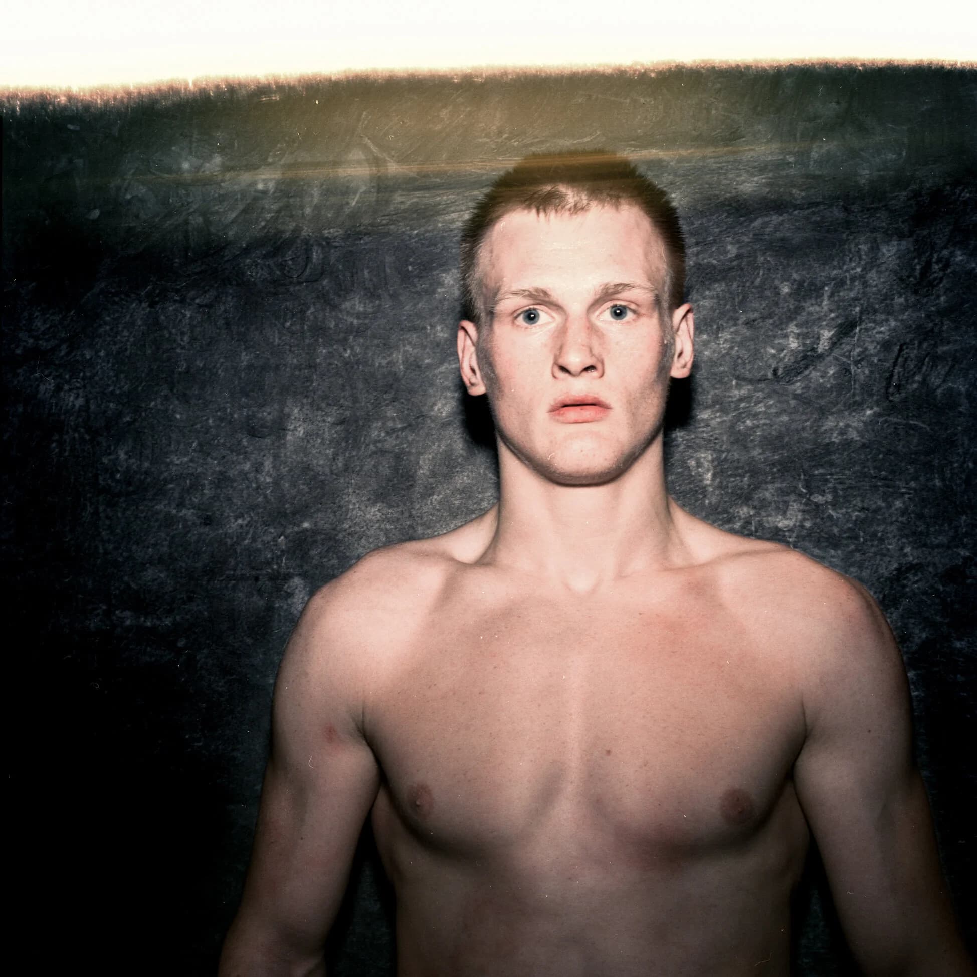 Post-fight portrait of a fighter with cropped hair and wide pale-blue eyes, flushed and dazed, light flaring at the top of the frame