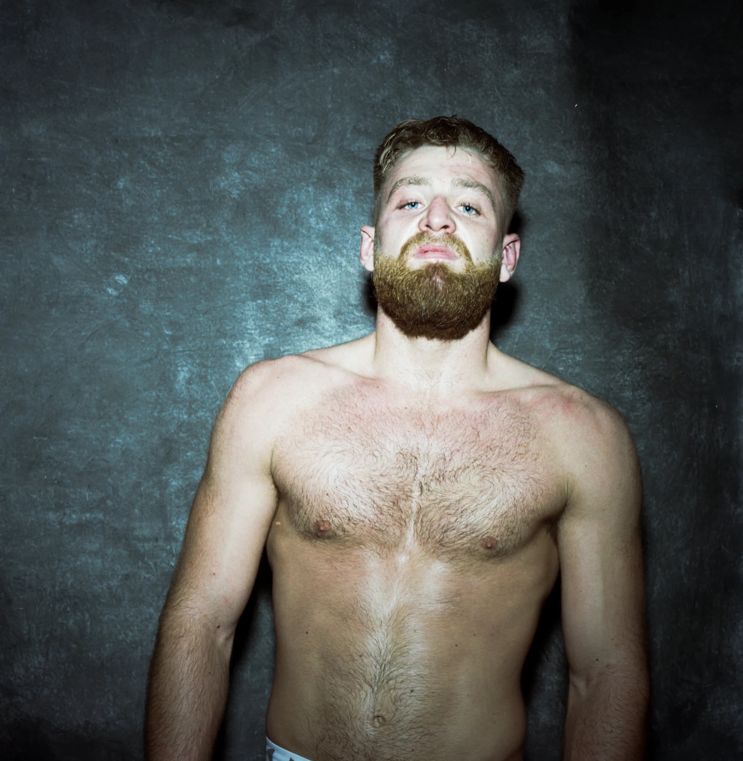 Post-fight portrait of a thick-bearded fighter with damp hair and chin raised, heavy-lidded gaze against a blue-grey backdrop