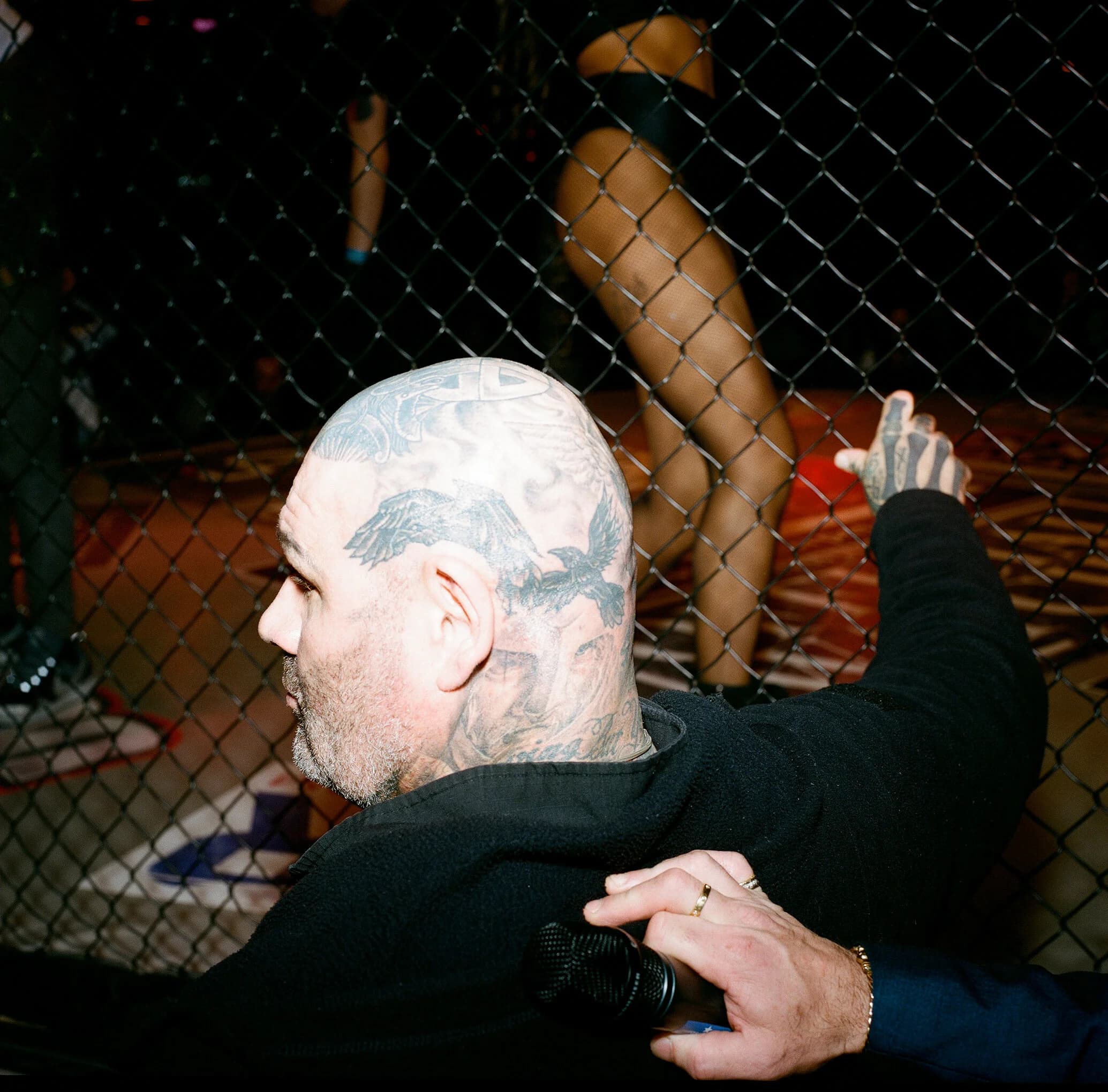 Bald man at cageside seen from behind, ravens tattooed across his scalp, holding a microphone as a ring girl's fishnet legs pass beyond the fence