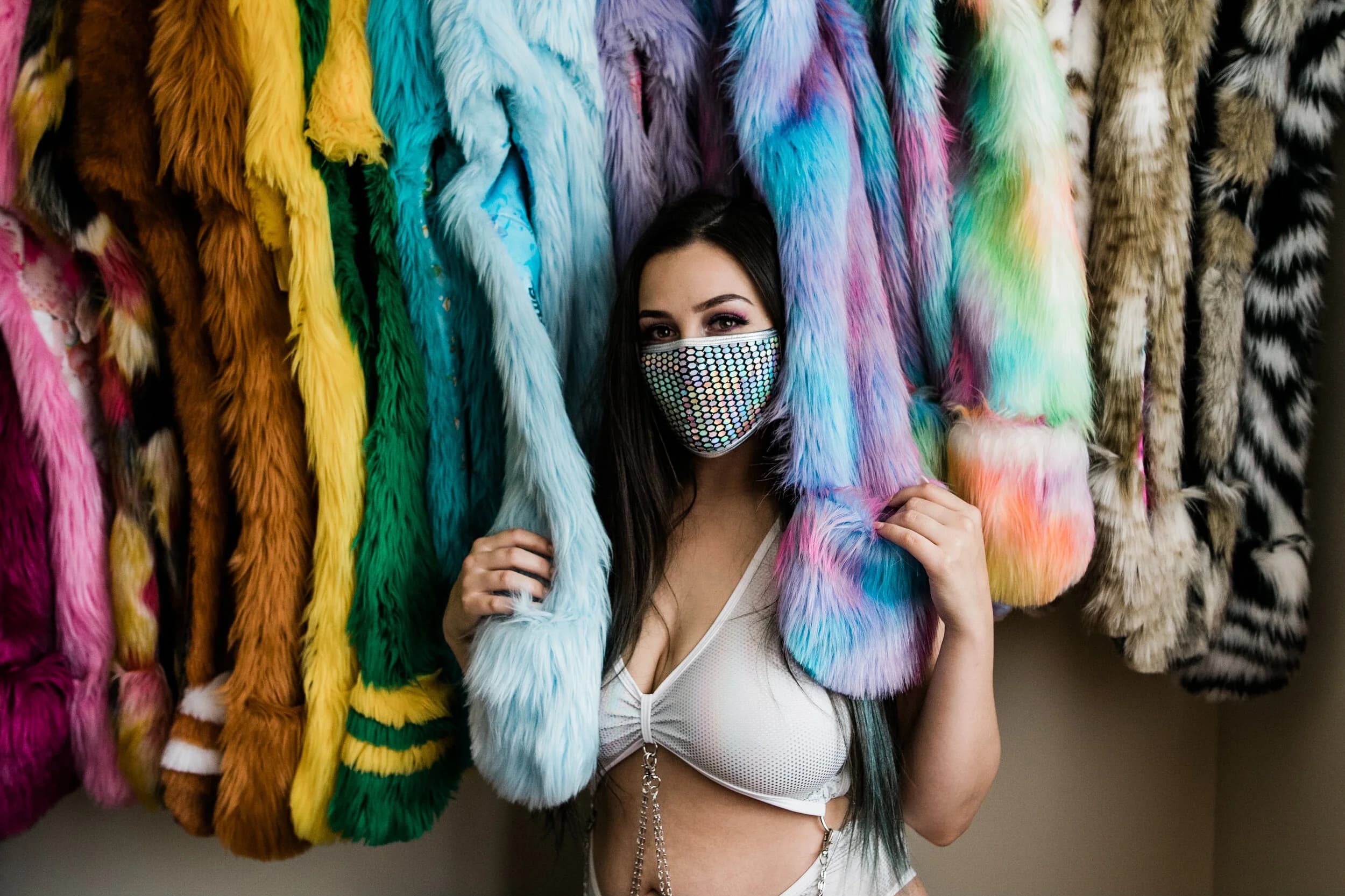 Raver in a holographic sequin face mask and white bra peering out between racks of rainbow faux-fur coats