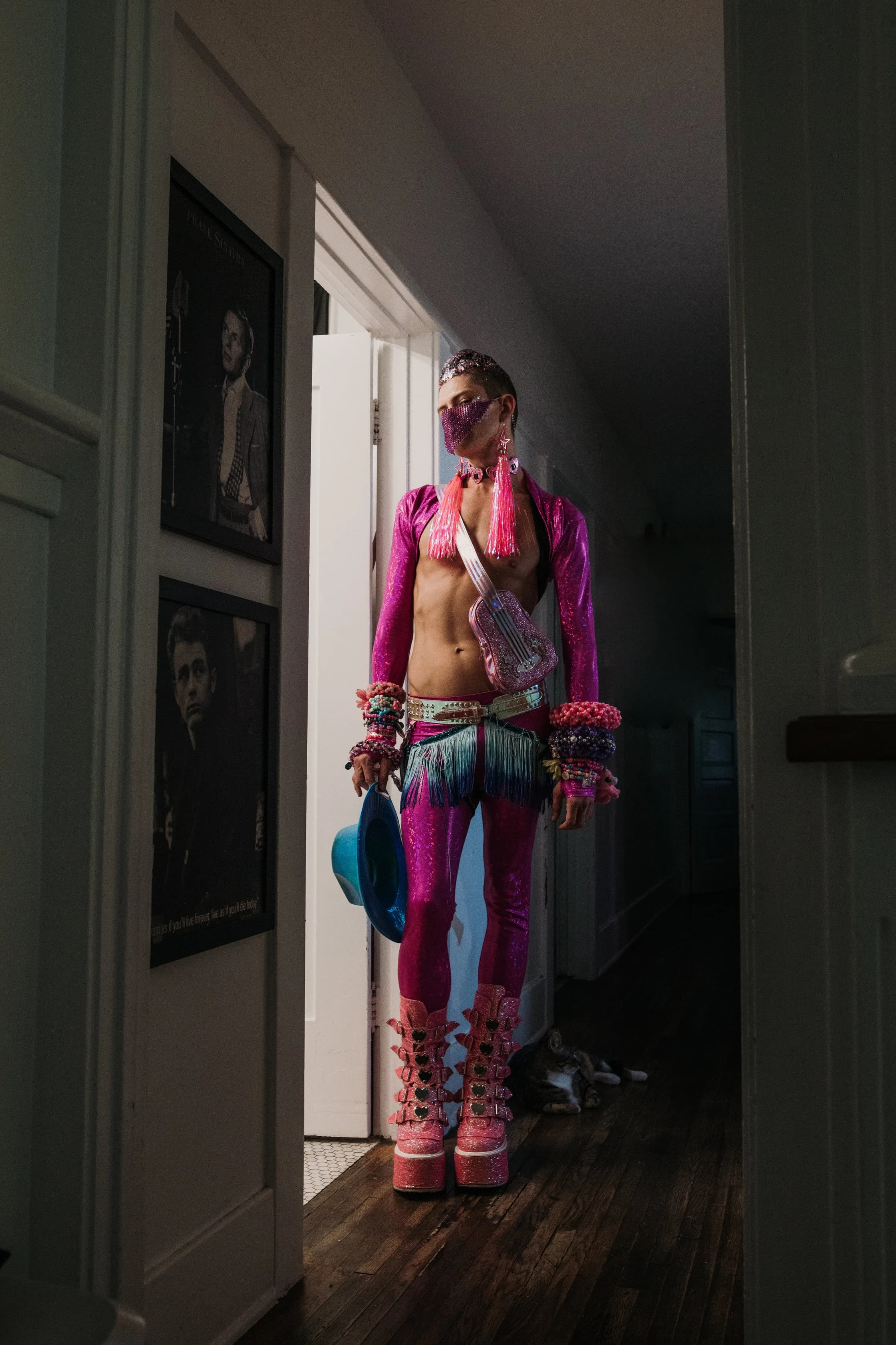 Raver in a metallic pink outfit with a rhinestone face veil, tassel earrings, stacks of kandi cuffs, a glitter guitar-shaped bag and pink platform boots, standing in a dark hallway doorway