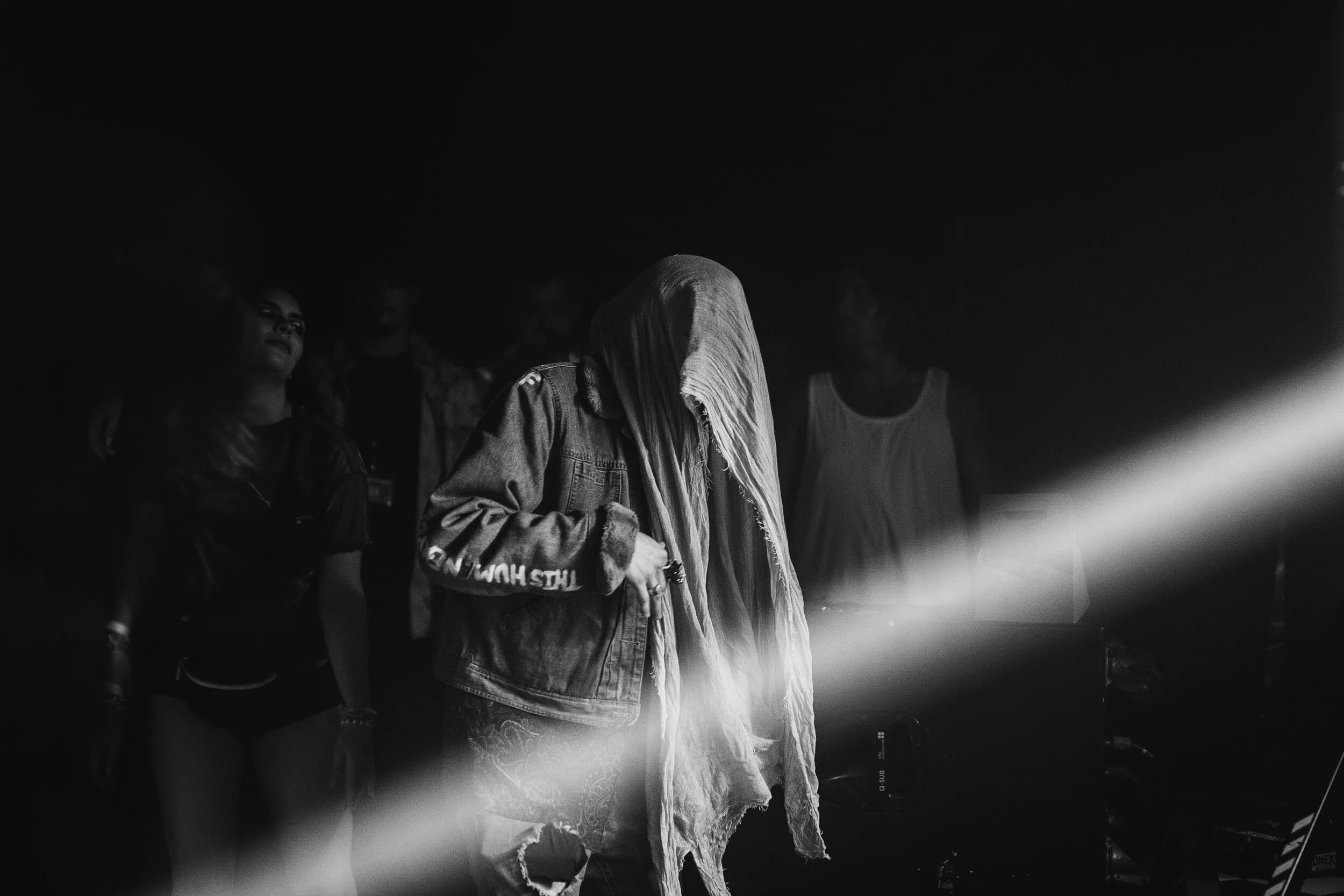 Black-and-white image of a dancer with a cloth draped fully over their face, wearing a denim jacket, a beam of light cutting across the dark crowd
