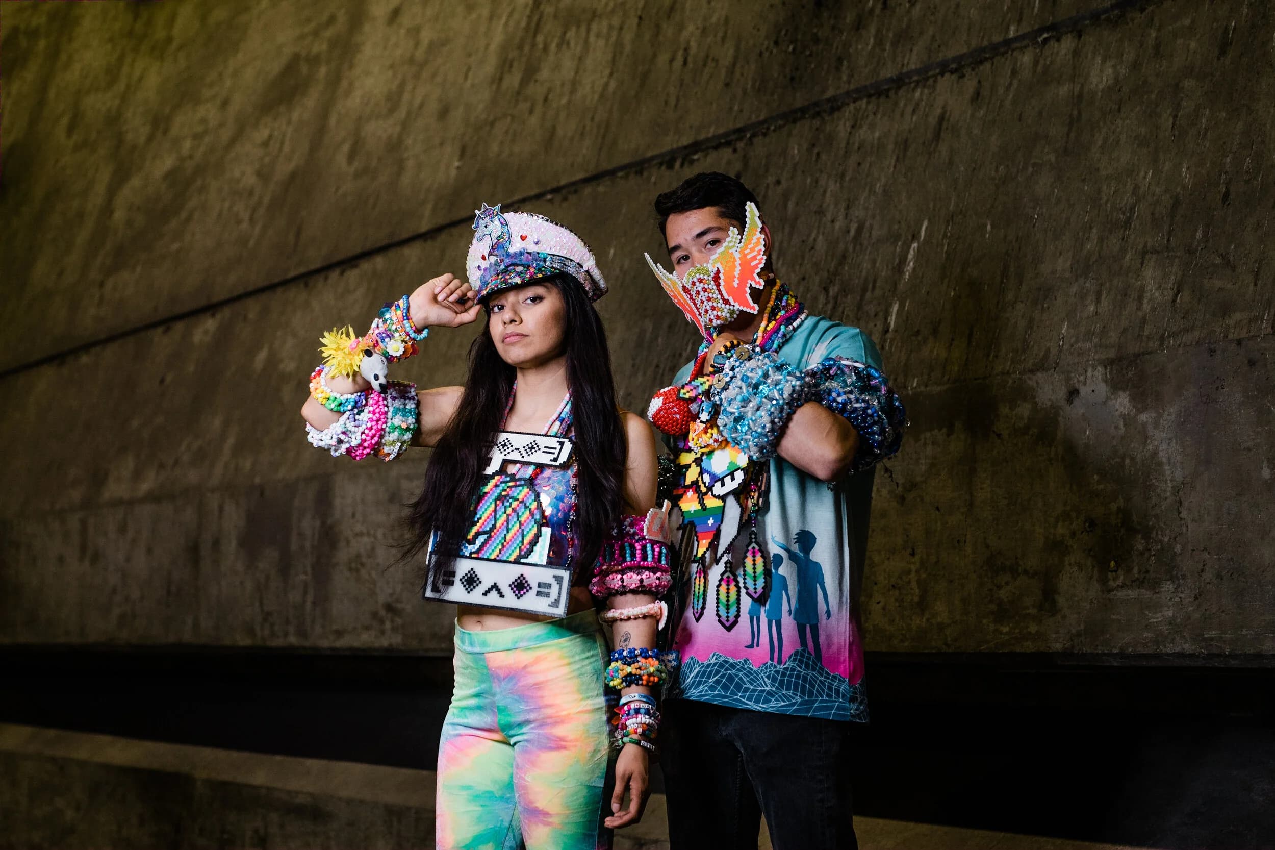 Two Kandi Kids posing in a concrete underpass, one in a bejewelled captain's hat and pixel-bead top, the other in a beaded pegasus face mask, both with forearms stacked in kandi