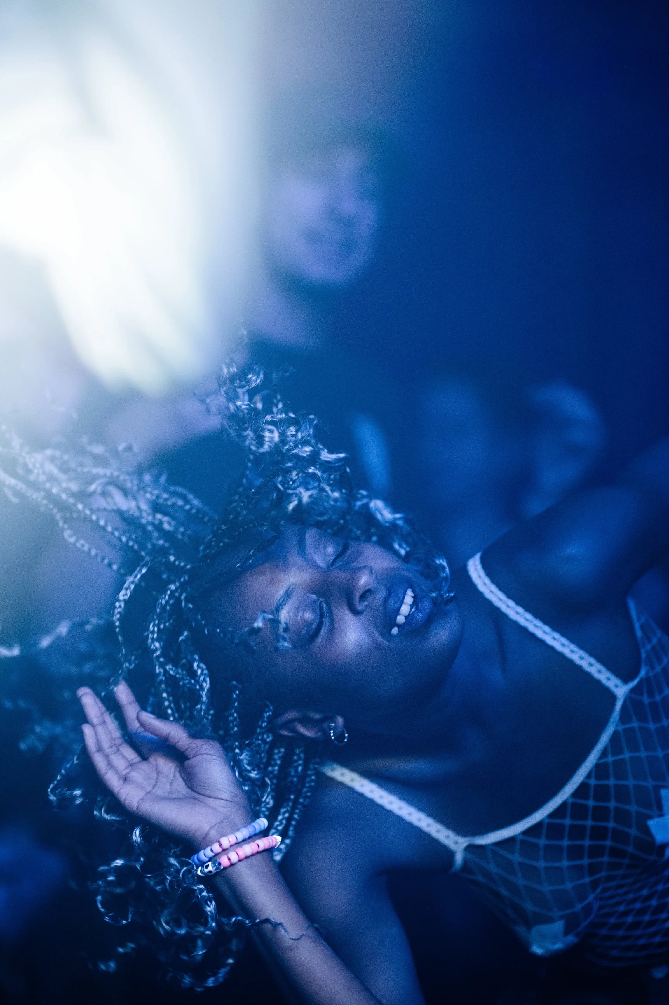 Blue-toned crowd moment as a woman with braids dances with her eyes closed, a kandi bracelet on her raised wrist