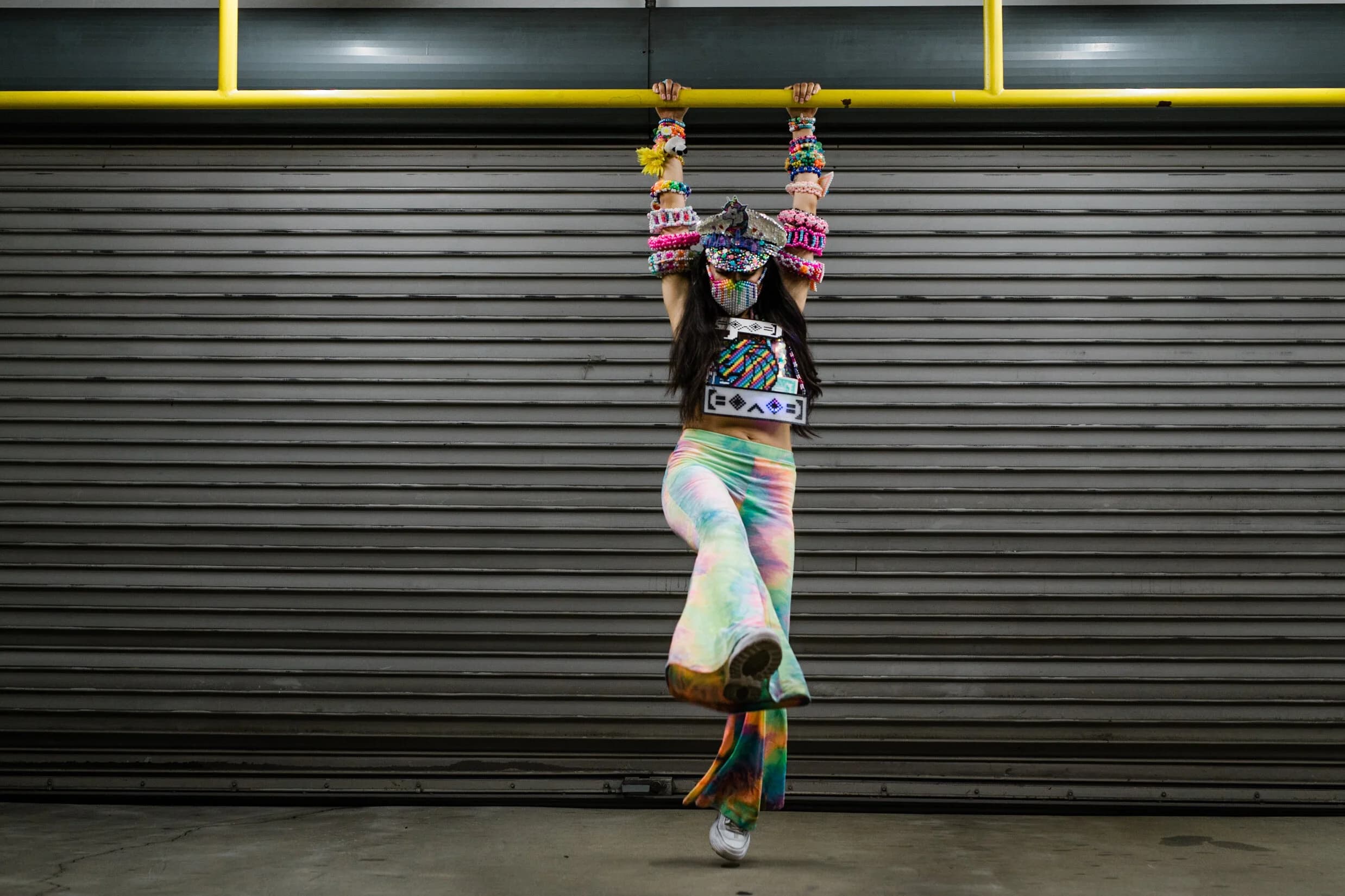 Raver in a unicorn captain's hat and beaded mask hanging from a yellow bar in front of a corrugated shutter, arms ringed in kandi and one tie-dye-flared leg kicked up