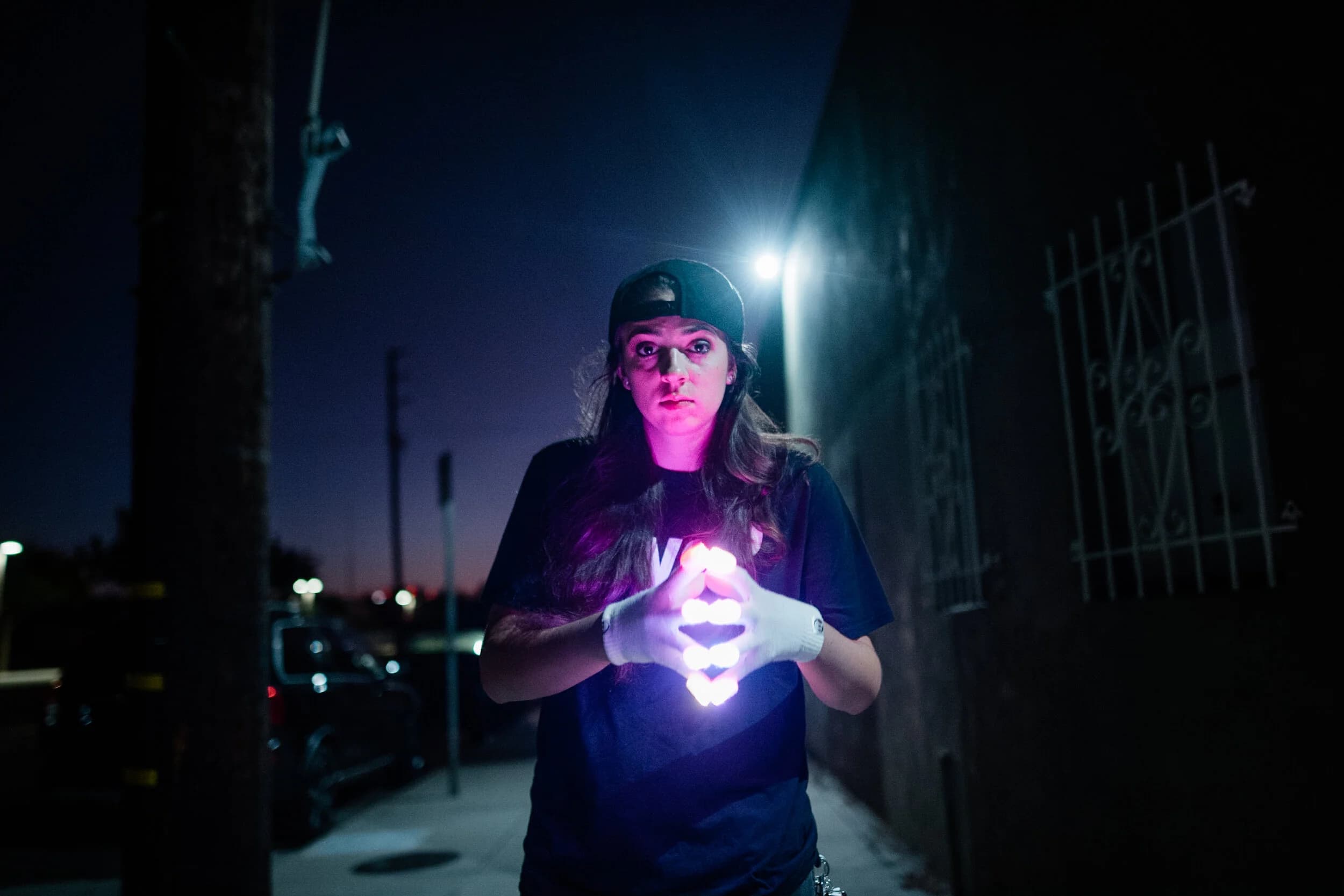 Woman in a backwards cap forming a glowing diamond with LED-gloved hands on a night street lit purple
