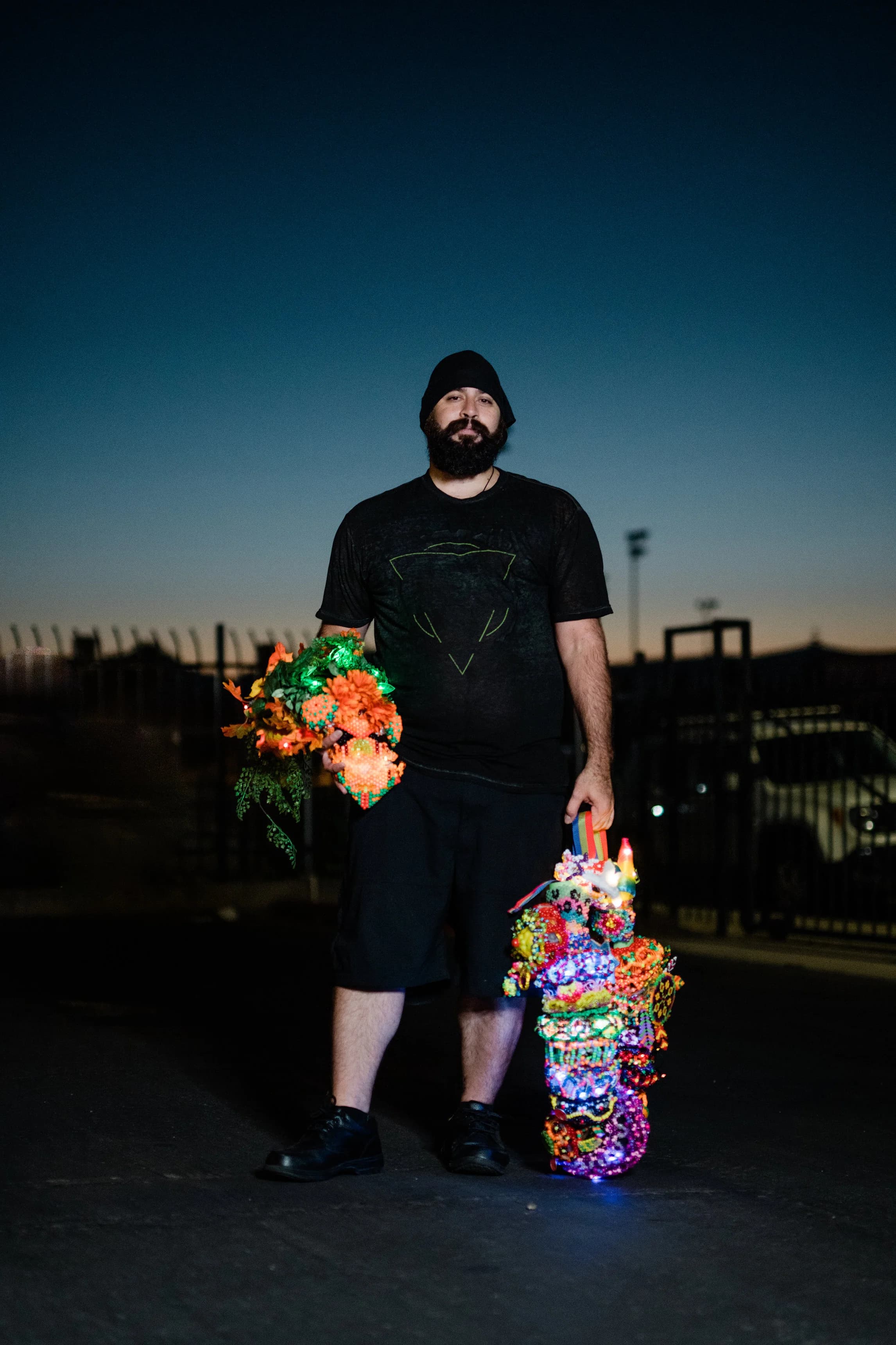 Bearded man in a beanie standing at dusk holding two LED-lit kandi totems, one a glowing flower bouquet and the other a stacked beaded staff