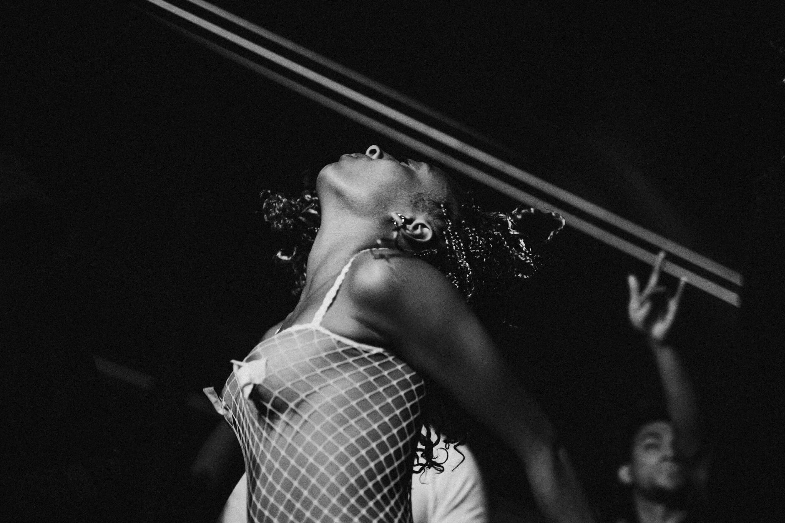 Black-and-white shot of a dancer in a fishnet top with her head thrown back beneath twin laser beams