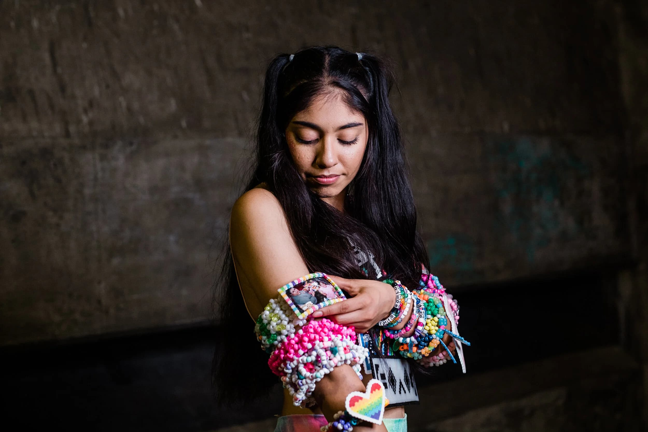 Raver with pigtails looking down at a photo kandi cuff on her arm, both forearms covered in beaded bracelets, a rainbow heart piece at her wrist