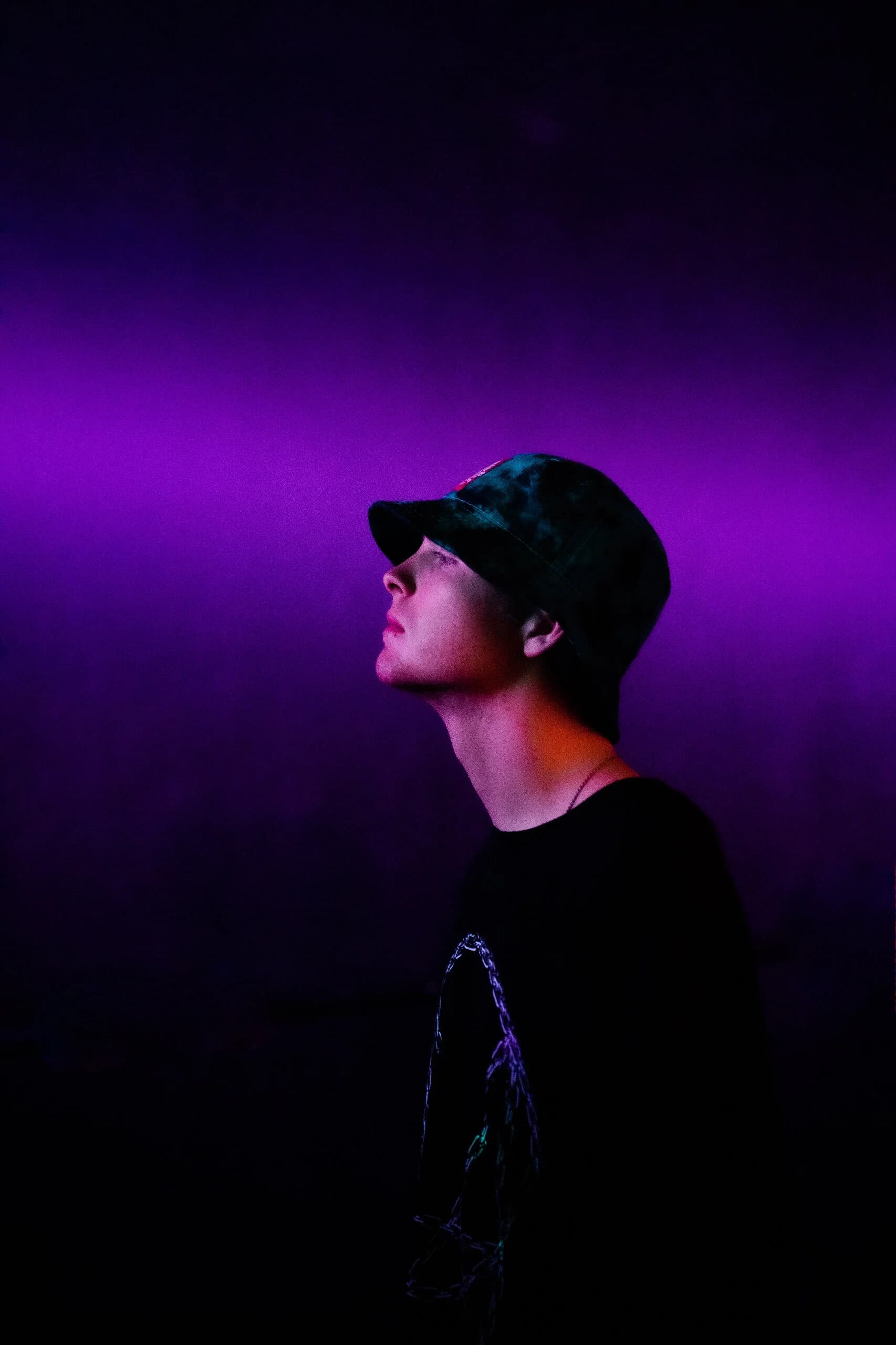 Profile of a young man in a dark bucket hat gazing upward against a saturated purple haze