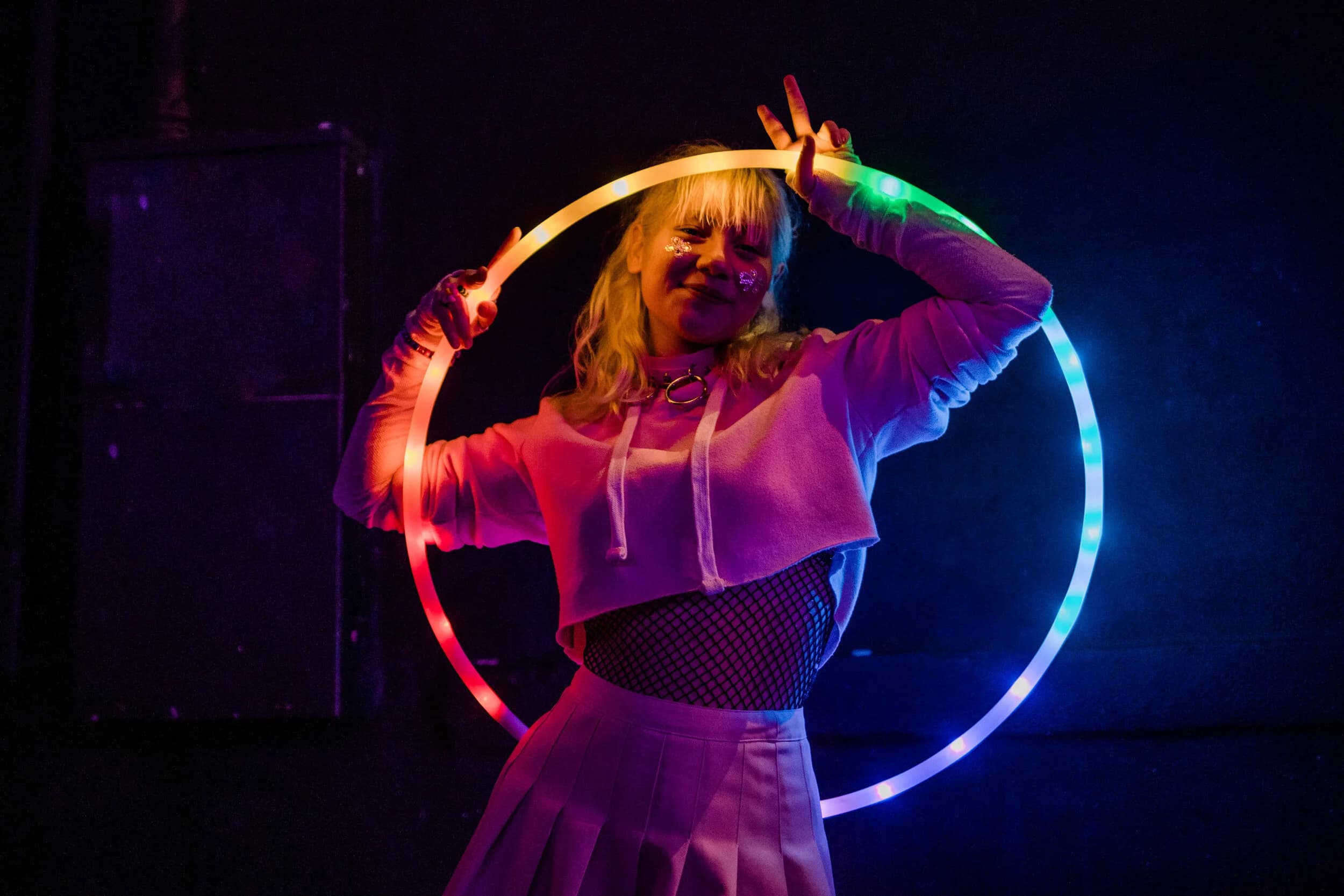 Blonde raver with glitter on her cheeks framed inside a rainbow LED hoop, flashing a peace sign